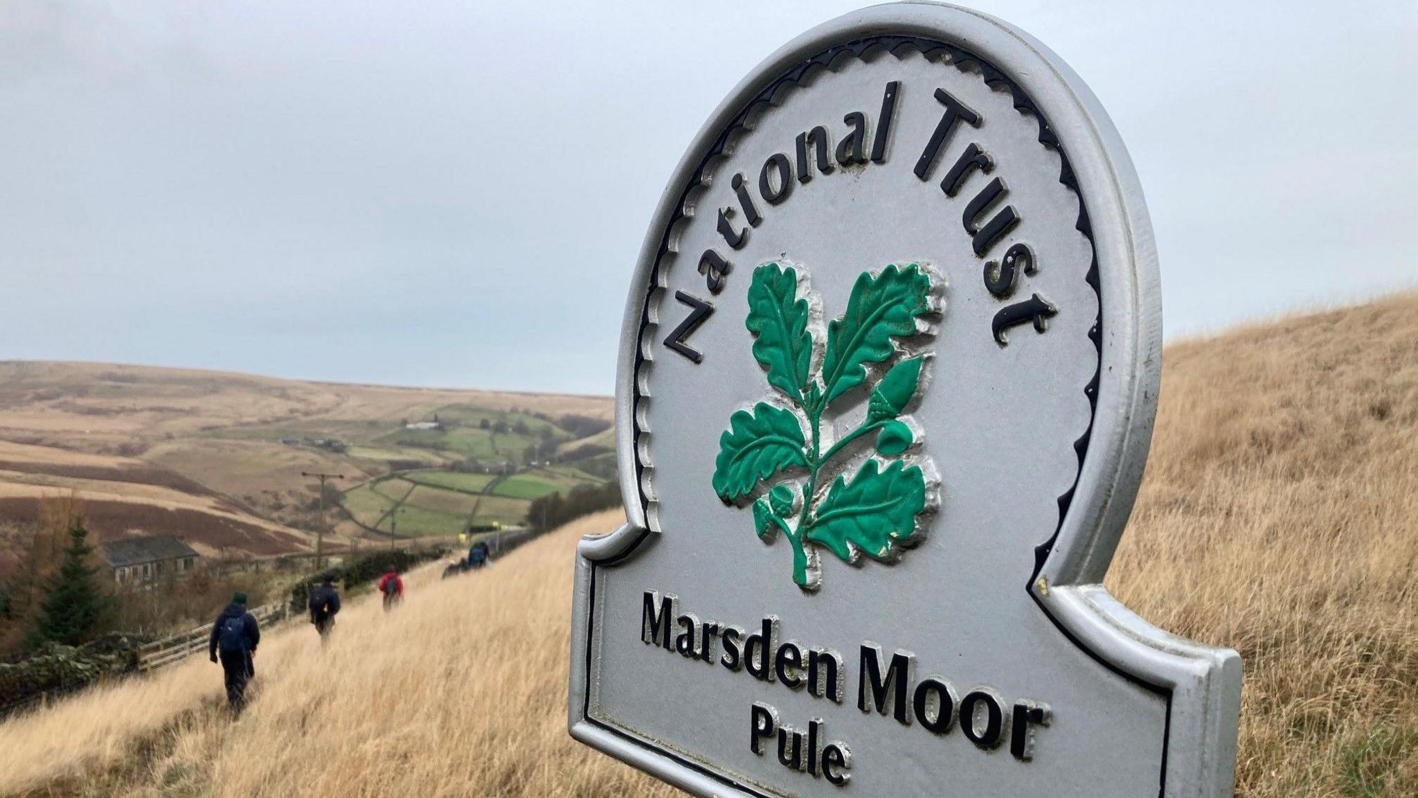 A cast-iron sign with the words Marsden Moor and Pule on the bottom and National Trust running around the top. There are some walkers in dark clothing in the background walking along a moorland path. 