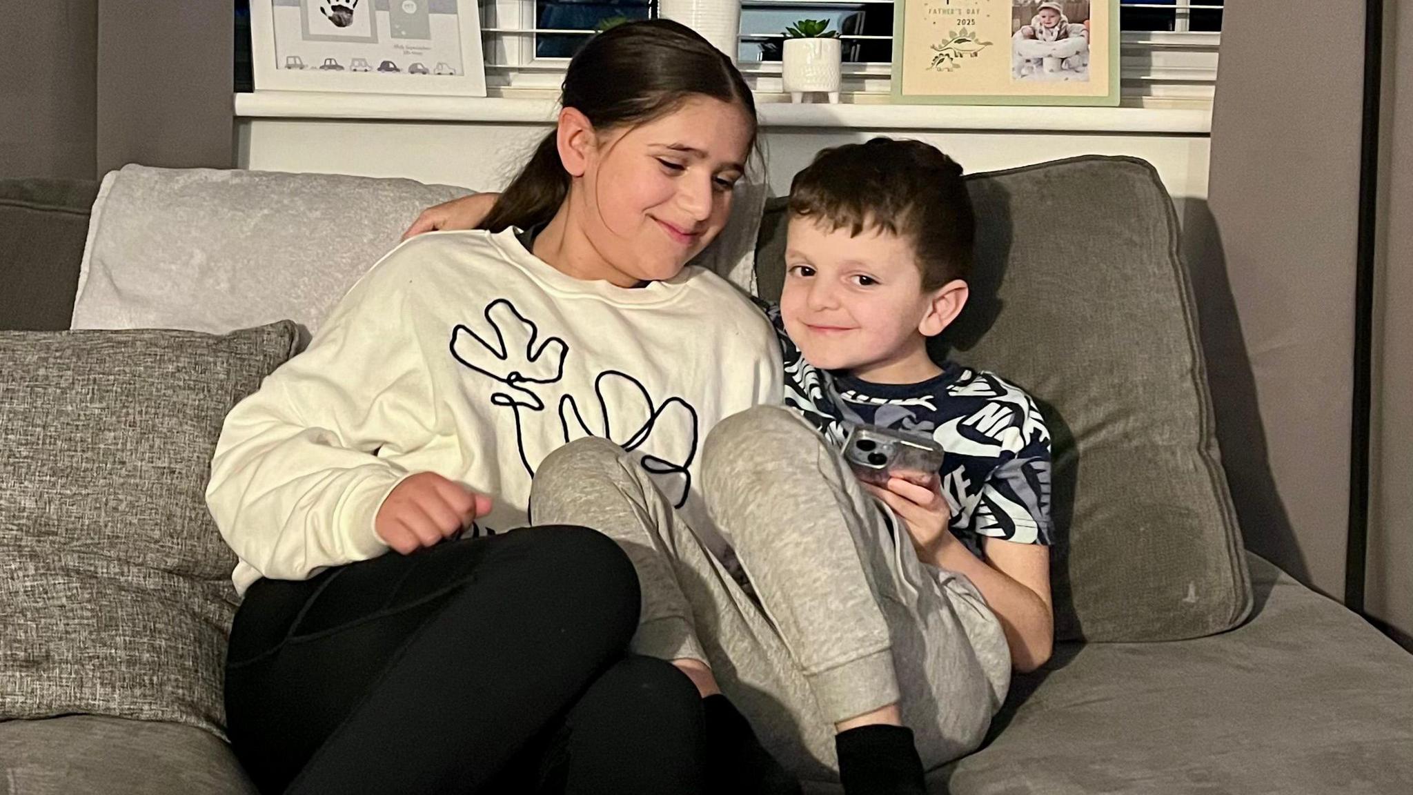 Olivia ac Arlo yn gwenu wrth eistedd gyda'i gilydd ar soffa - mae braich Arlo o amgylch ysgwydd ei chwaer fawr