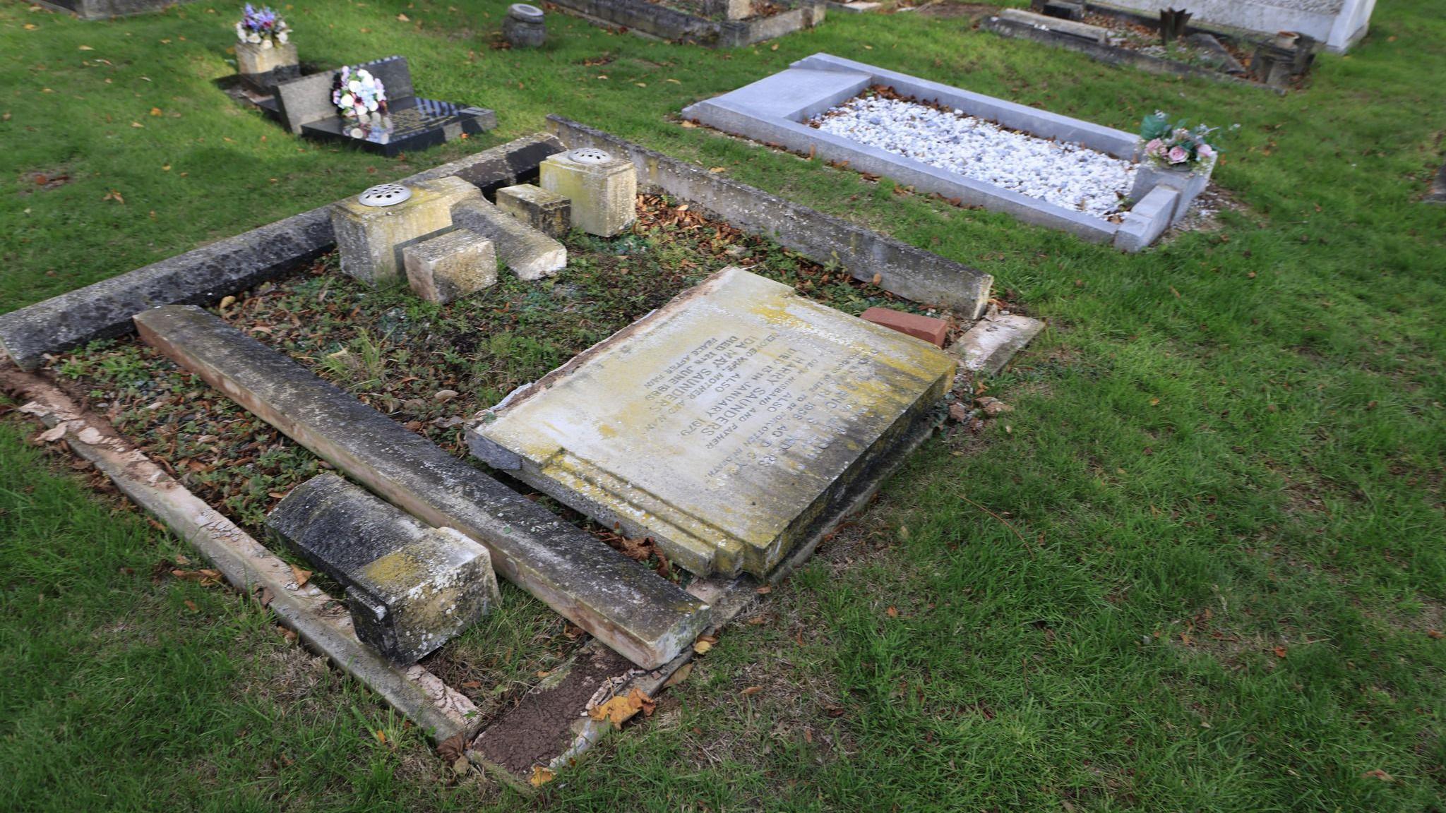 A stone headstone lies on the ground. Another headstone can be seen lying on the ground behind it.
