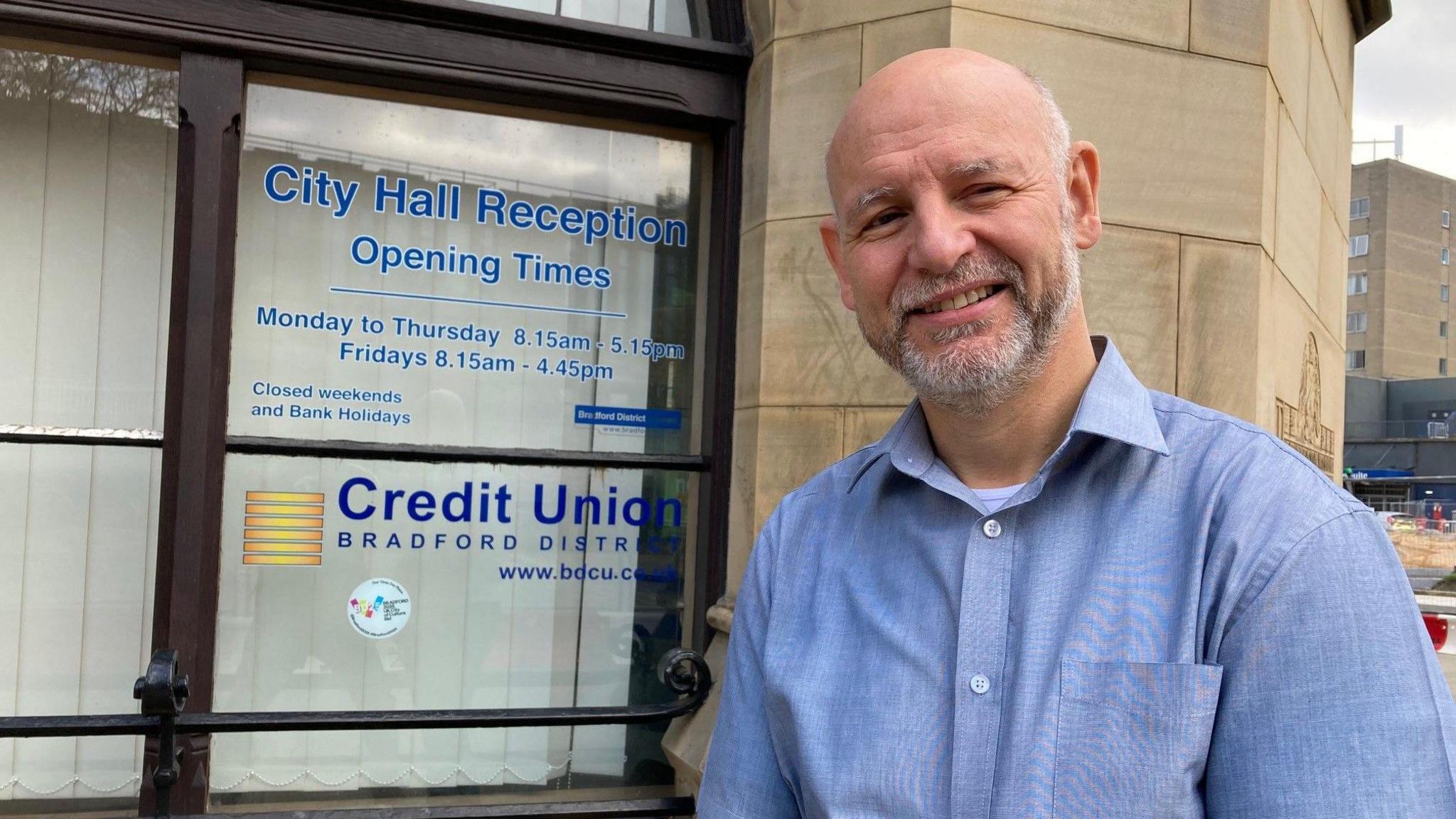 A bald man with a white beard and wearing a blue shirt stands smiling outside a window which has the details of the Bradford District Credit Union  on a see-through display.