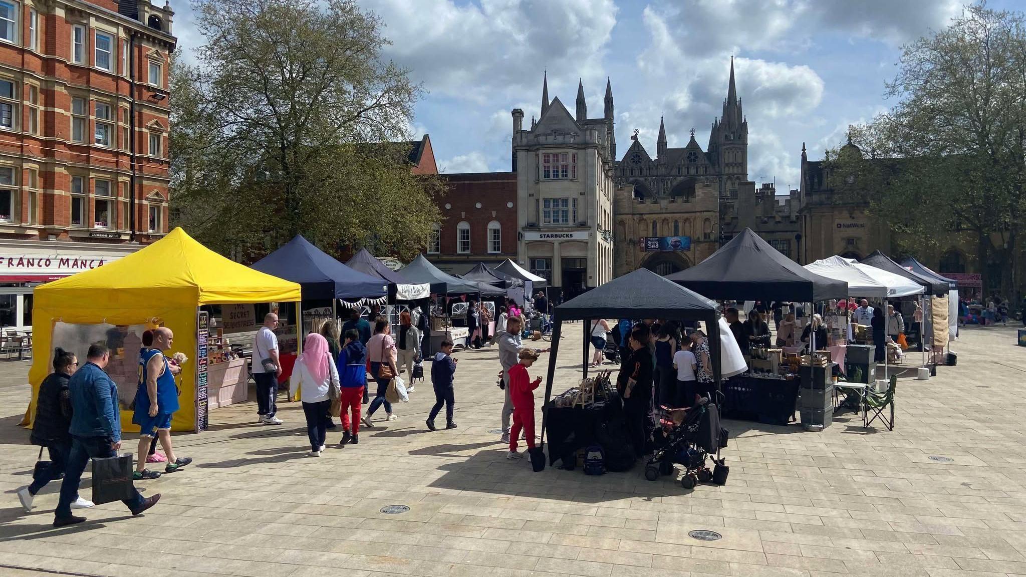 A food fair with yellow, black and white canopy stalls in a town centre square. People are walking around and visiting the stalls.