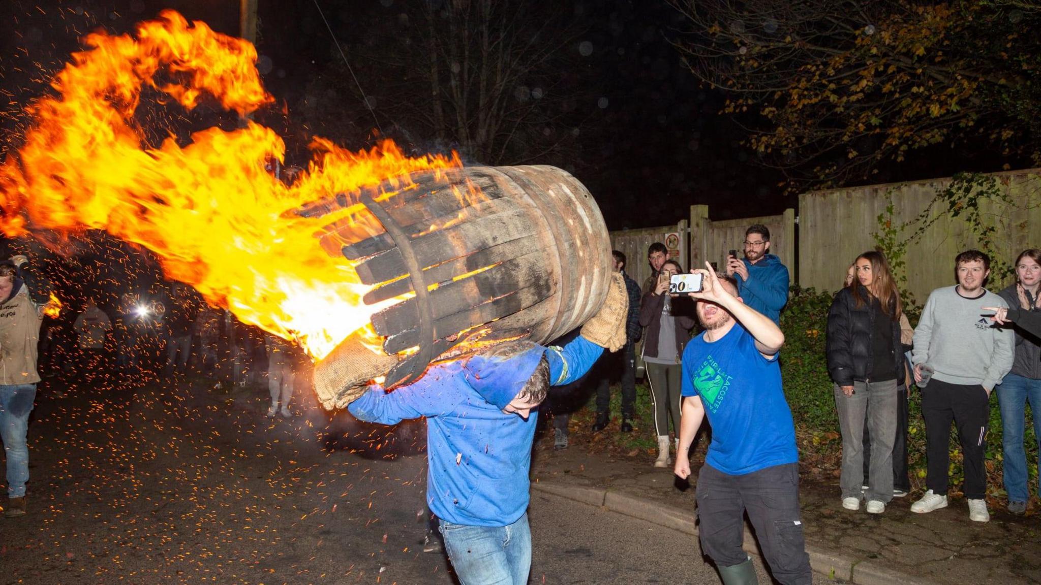 A man wearing a blue hooded top walking down a street with a flaming barrel on his back