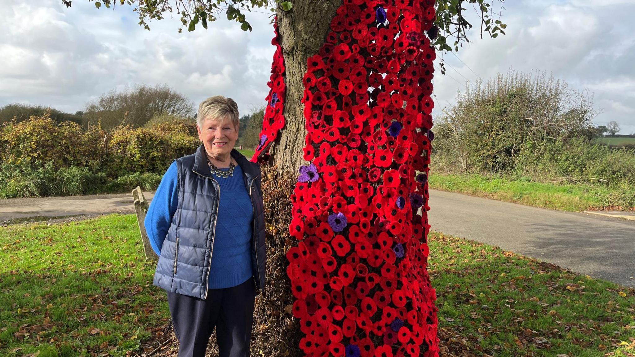 Woman stands in front of a tree which is covered in a cascade of knitted poppies in various shades of red and purple. The woman is smiling and wears a navy blue gilet over a royal blue jumper and a necklace.