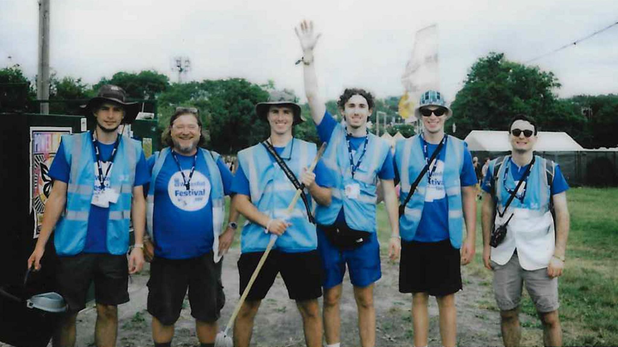 A film photo showing a group of six WaterAid volunteers wearing hi-vis branded blue vests and lanyards. They are carrying equipment like a mop and a bucket to clean toilets. They are smiling and posing for a photo.