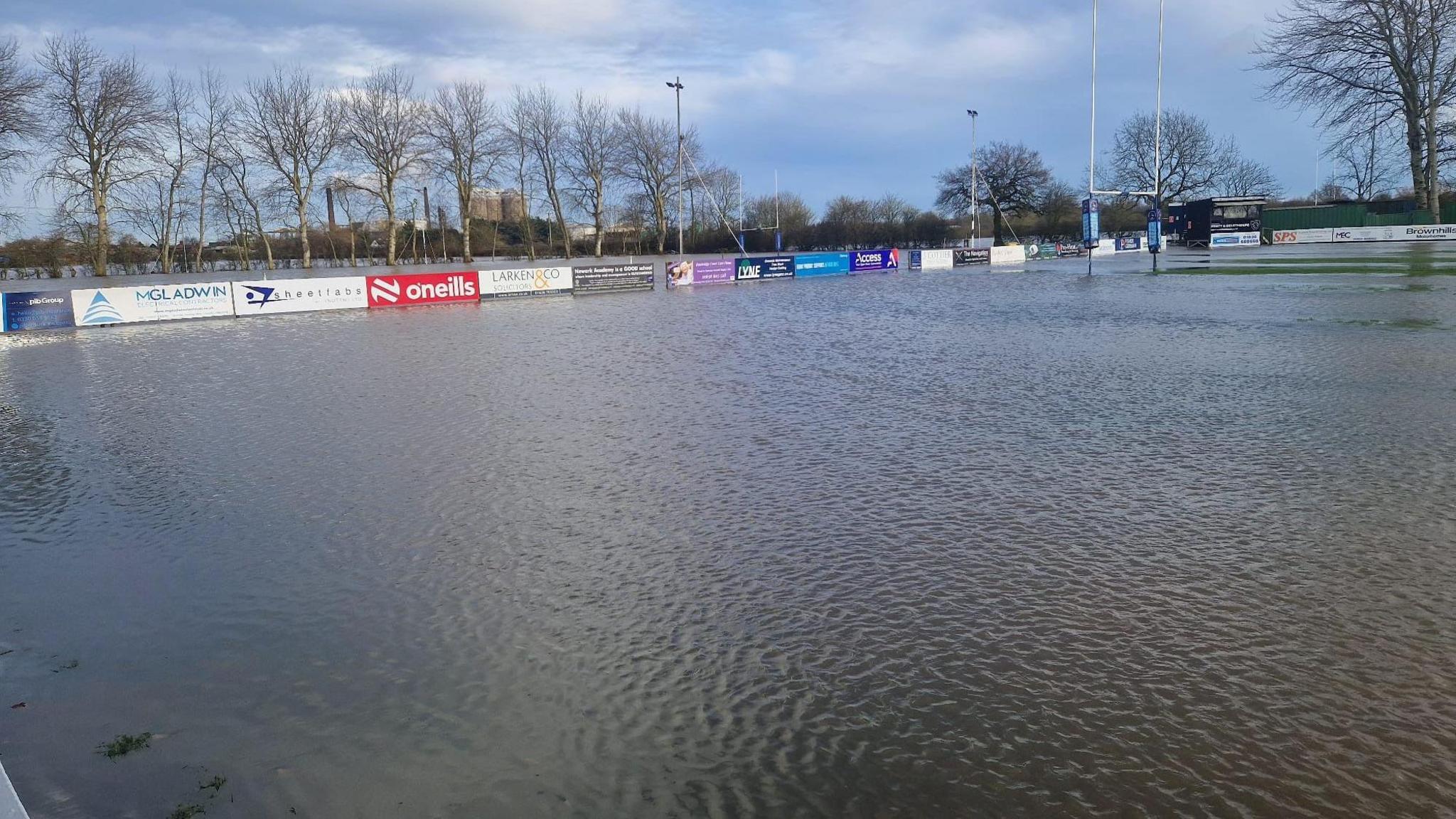 Wide shot of rugby pitches completely submerged, with goal posts and advertising boards the only items visible. Trees are in the background. 