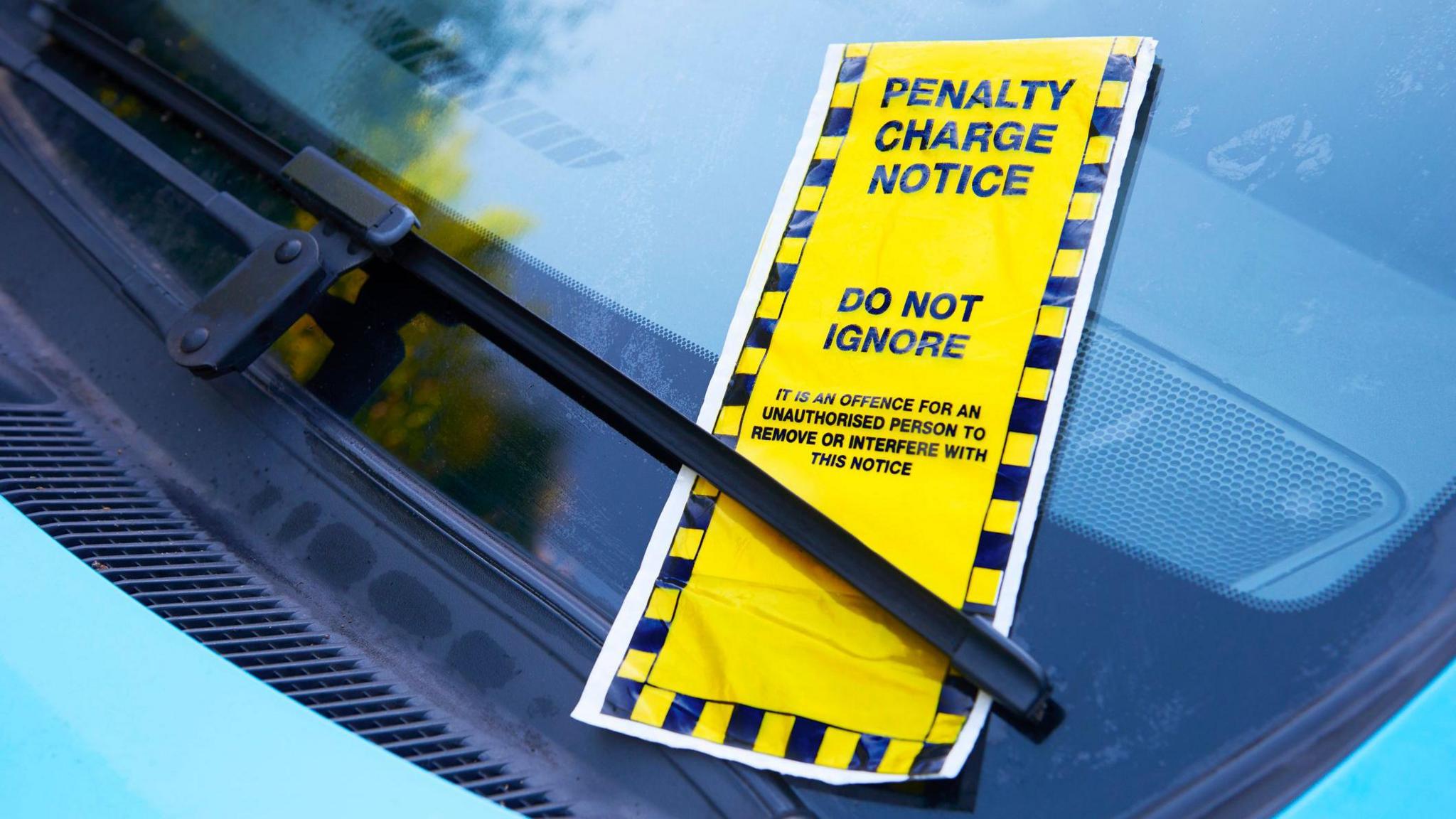 A yellow parking ticket which reads "do not ignore" slipped under a windscreen wiper of a car.