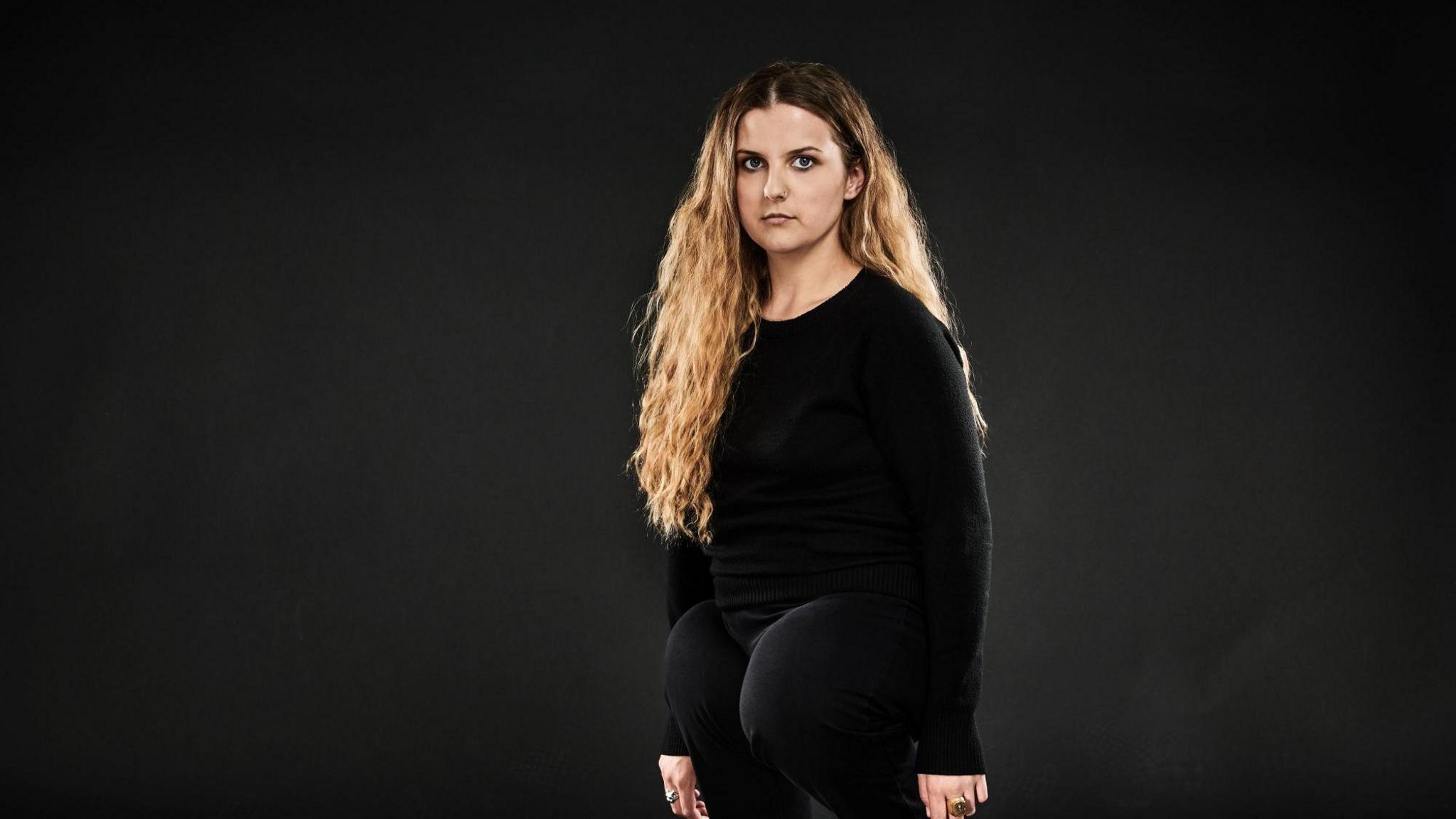 Ella Glendining wearing a black shirt and jeans stands and looks at the camera. She has long blonde hair. Ella has a congenital, rare leg disability.