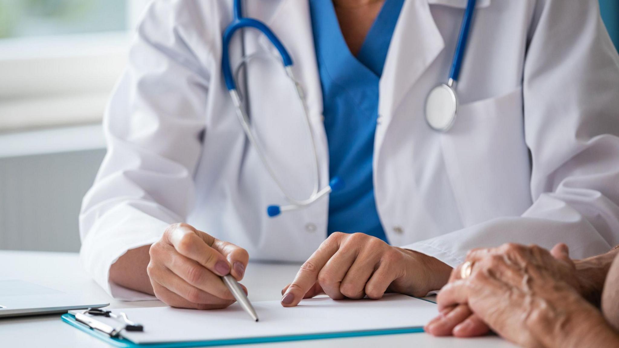 A doctor in a white coat with a blue top underneath, and a blue stethoscope around their neck. They have a pen in one hand and are pointing at a clipboard on a table with the other. A patient is sitting next to them with their hands clasped