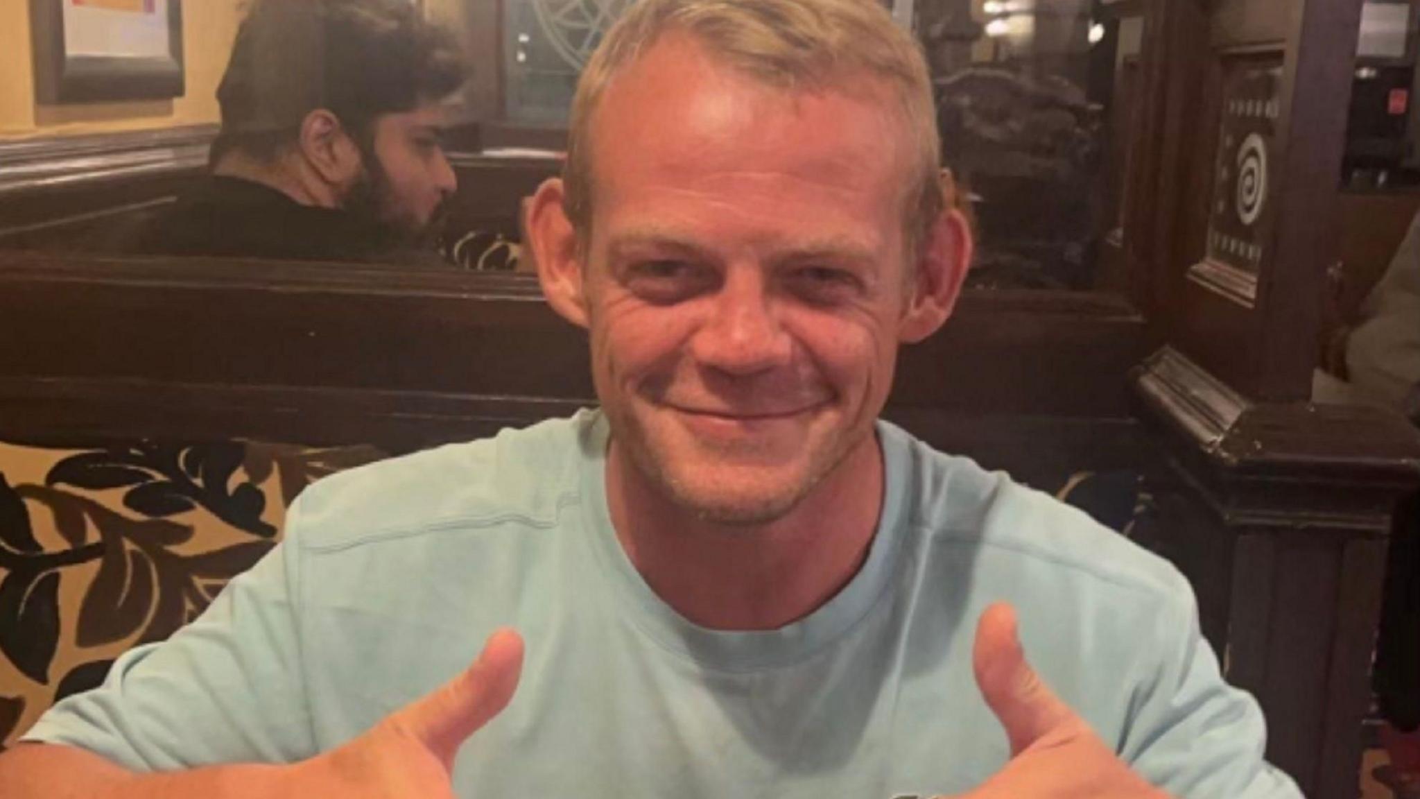 Andrew Main, wearing a blue T-shirt, sitting in a pub and giving a double thumbs up