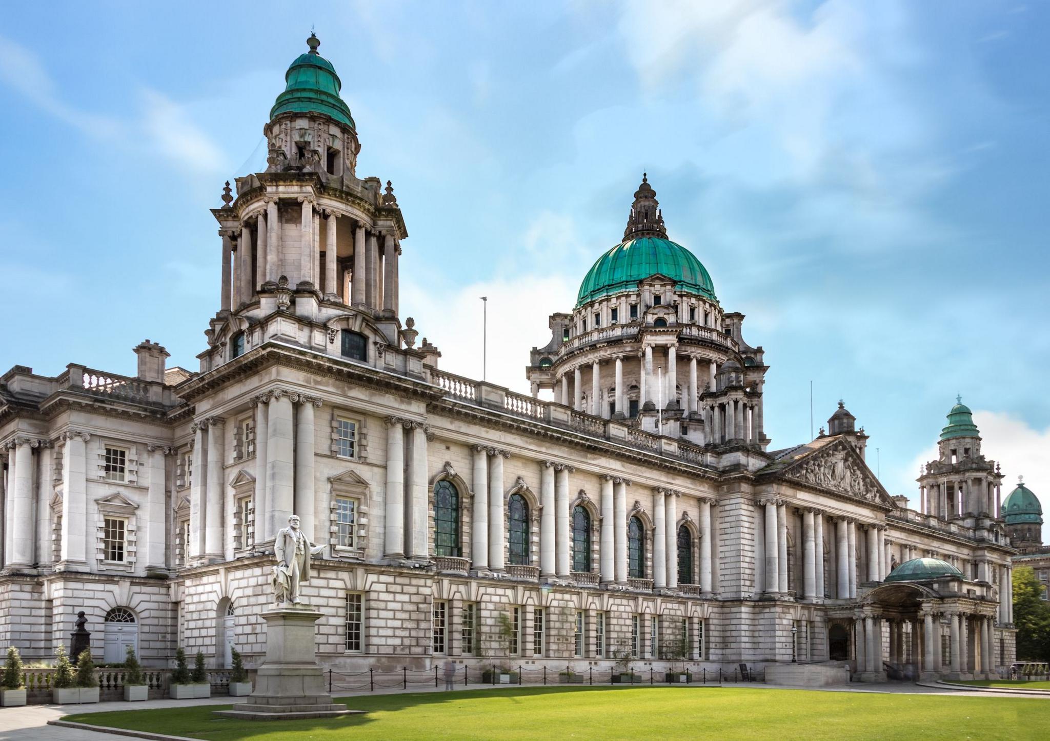 City hall is a large white turreted building with green roofs on the turrets. A large green lawn is in front of the building and the picture is taken on a bright day with blue skies.