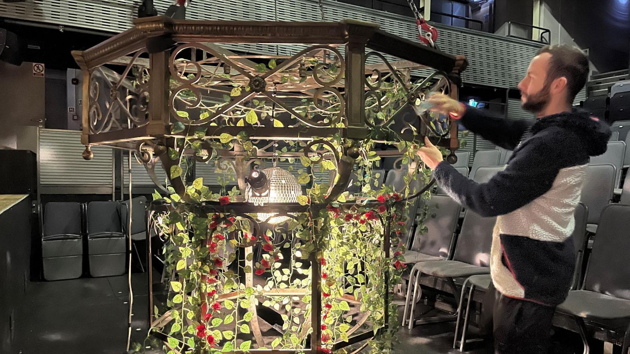 A chandelier light fitting draped in vines and roses. A man wearing a black and white top and black trousers is stood at the side making adjustments to the chandelier