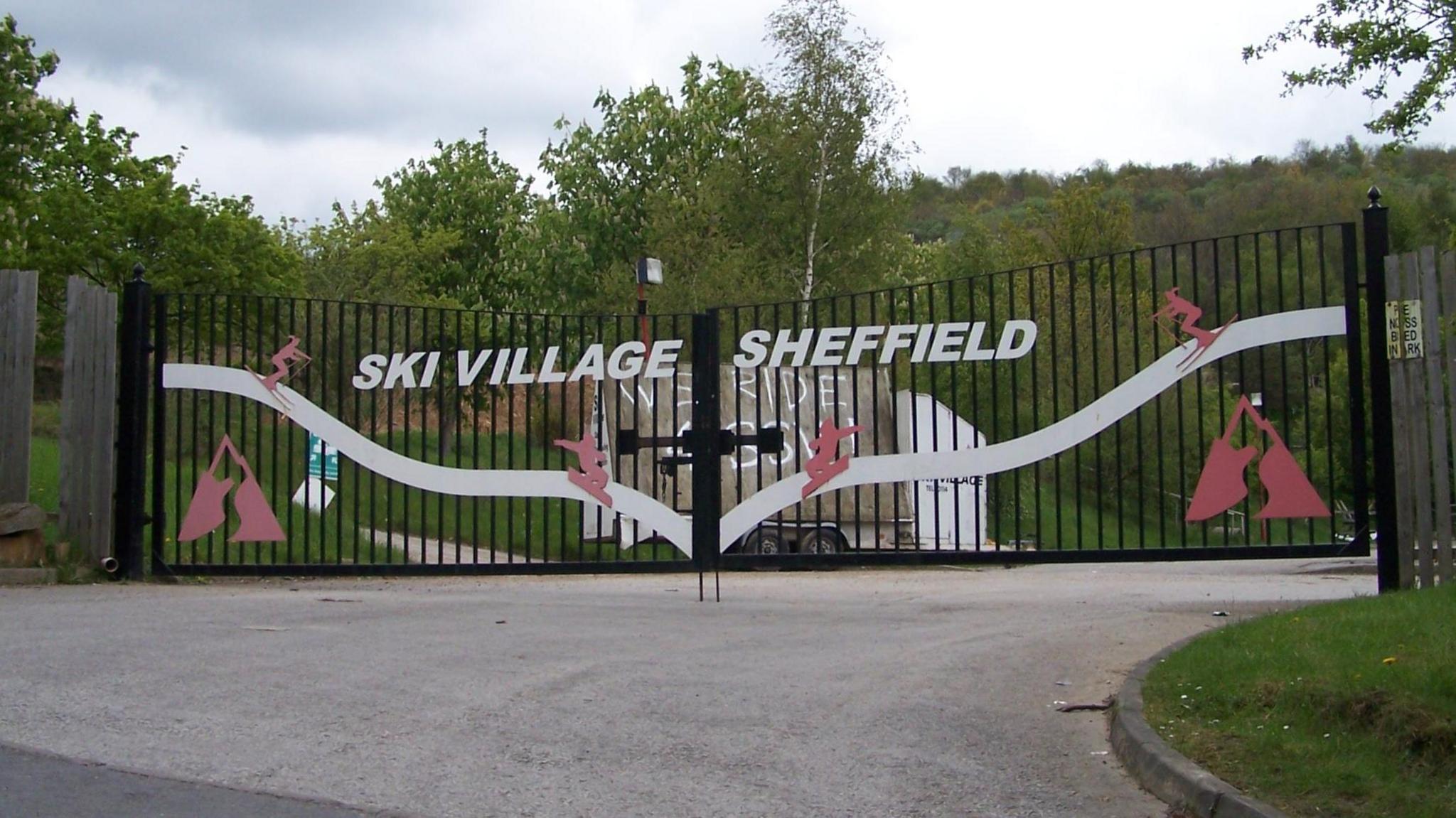 Large black gates with green trees and hills behind. On the gate it reads Ski Village Sheffield.