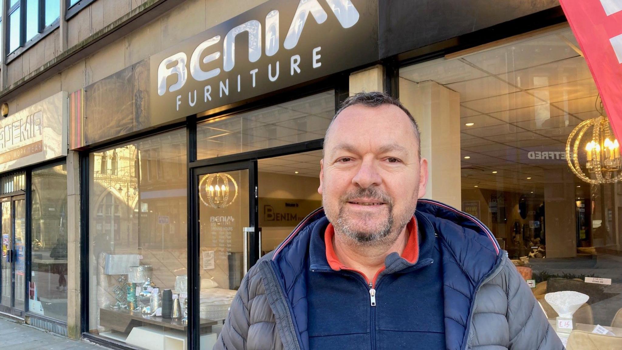 A man with grey goatee wearing a grey and blue jacket standing outside a shop front.