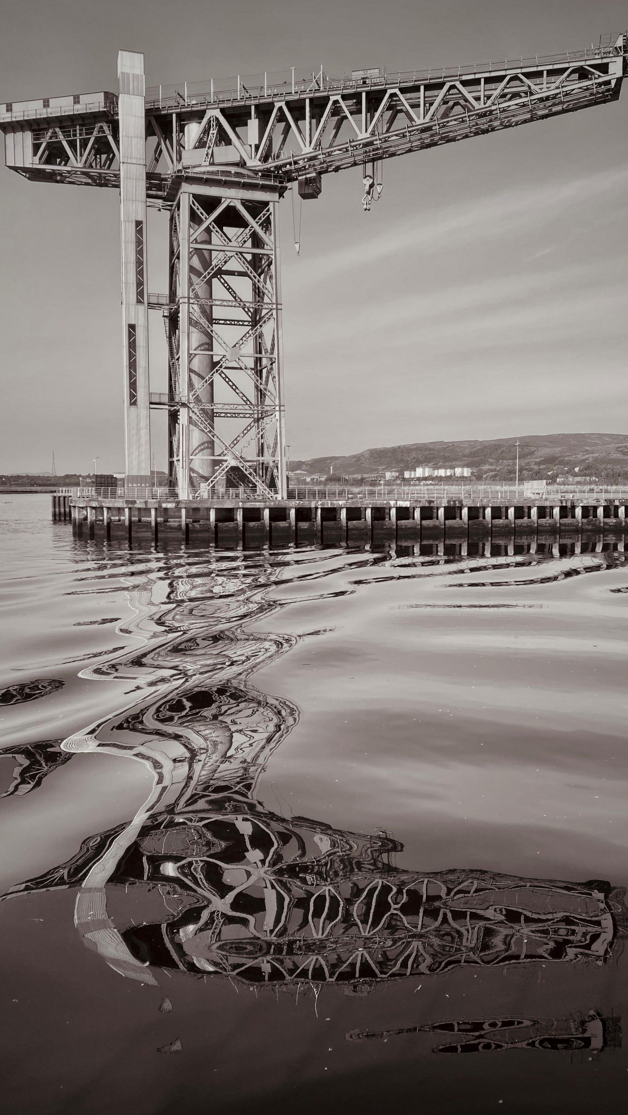 Black and white image of a crane, taken from below from a boat in the water next to it. The reflection of the crane is distorted in the water by light rippes.