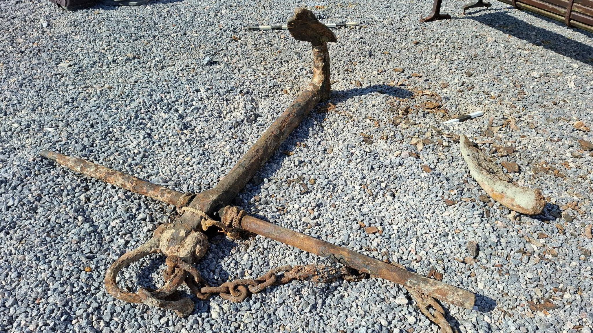 The anchor photographed on land on pebbles. One of its arms appears to be missing. It appears to be near a bench. It's a sunny day.