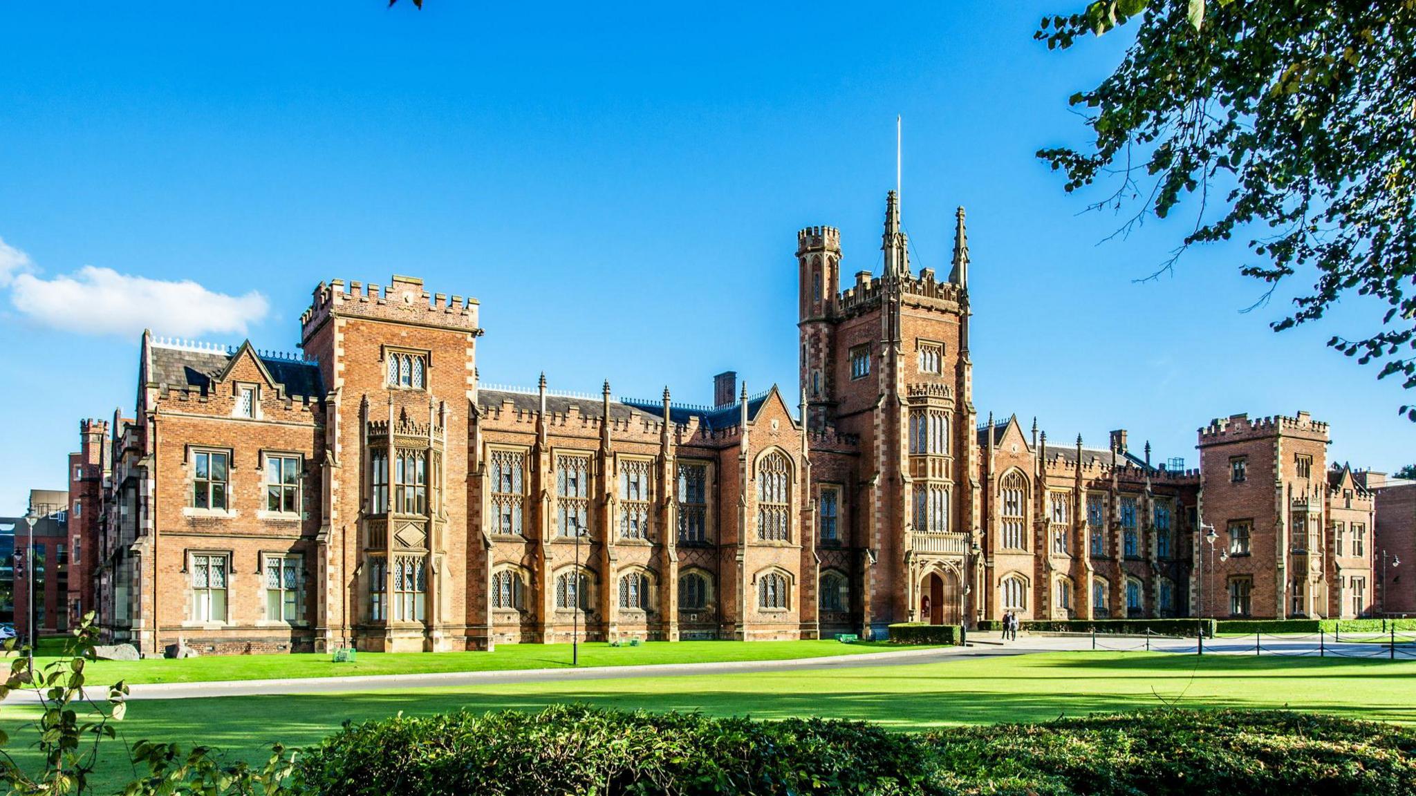 The outside of Queen's University Belfast. There is a large area of grass in front of the building and a blue sky above. 
