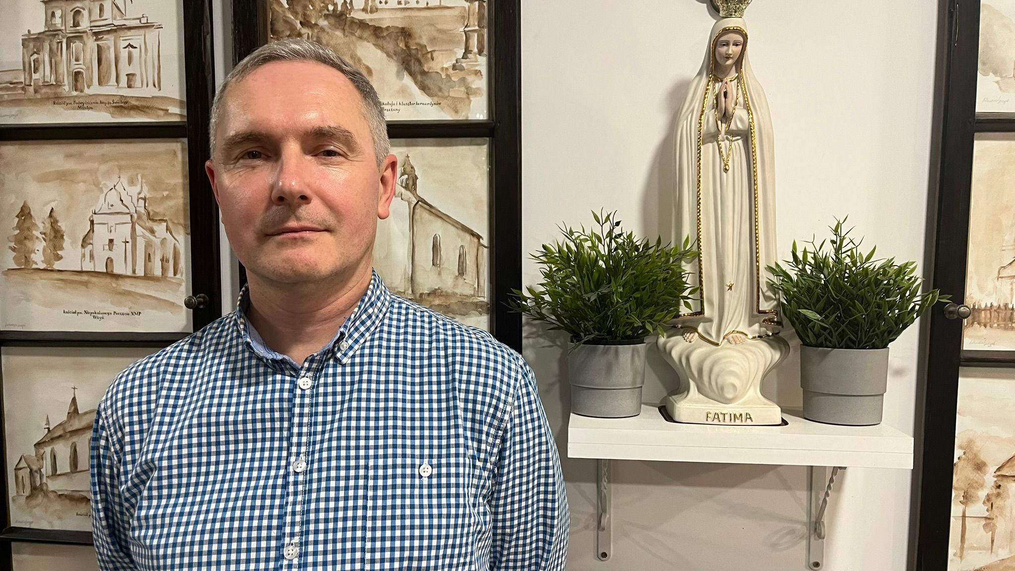 A man with short grey hair in a white and blue checked shirt stands in front of a series of paintings of churches, a statue and some potted plants 