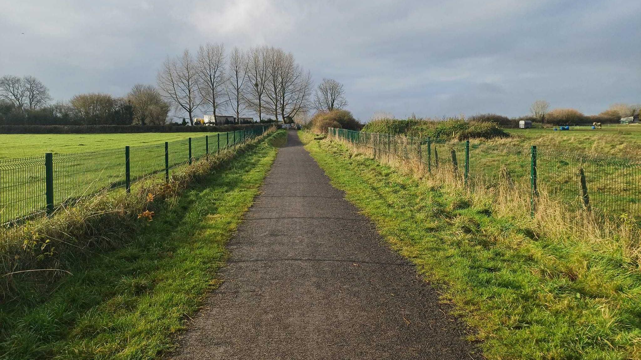 The Strawberry Line footpath near Ridge Road in Shepton Mallet. It is a wide tarmacked path with a metal fence lining either side, behind which are green fields and trees.