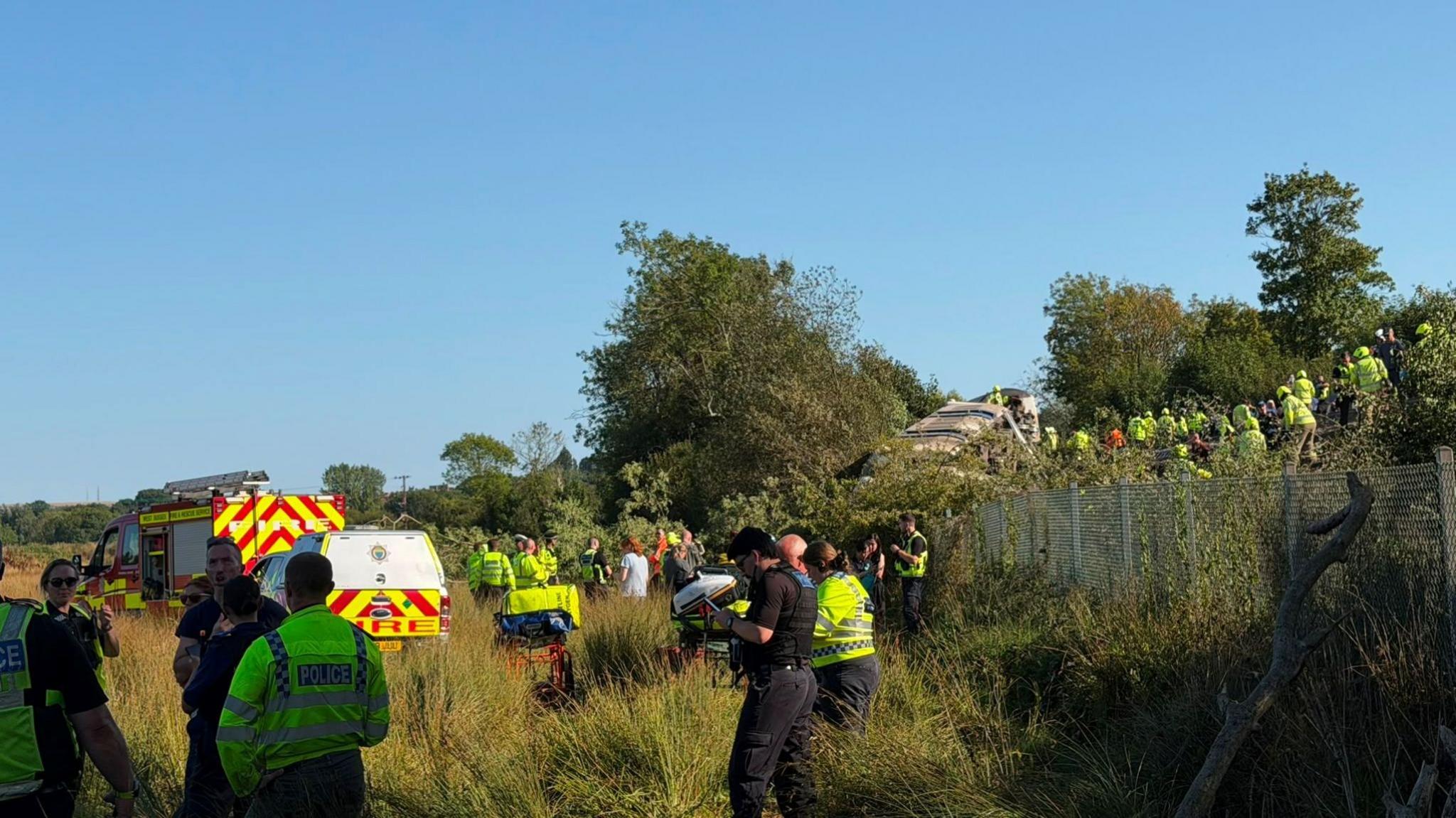 Emergency service vehicles, police officers and rail staff gather beside a railway embankment following a train derailment. People can be seen on and around the railway line above, while responders work in a grassy field below.