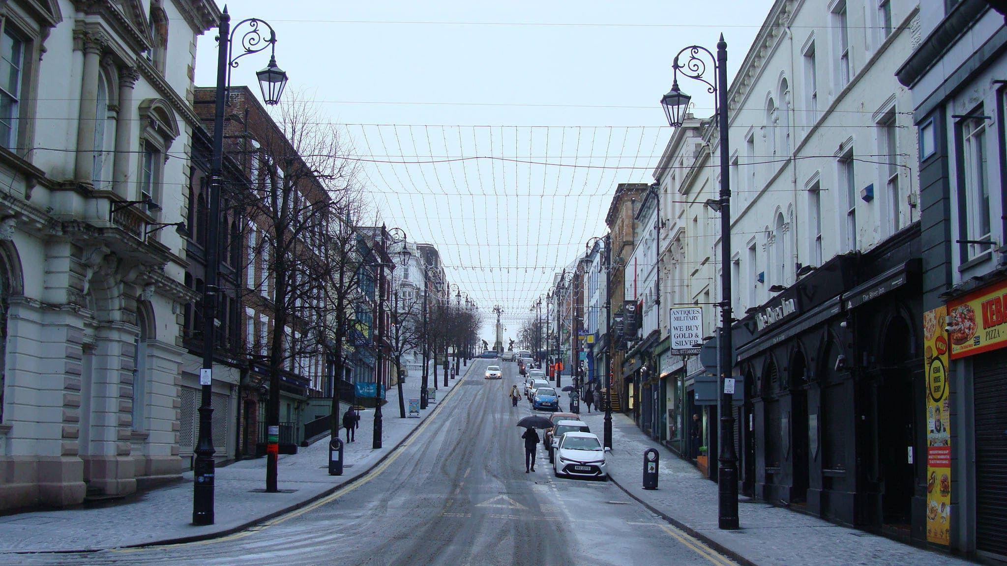 An icy Shipquay Street in Derry, cars are parked on wither side of the steep street, with a single pedestrian walking across the road. ice is clearly visible on the road.