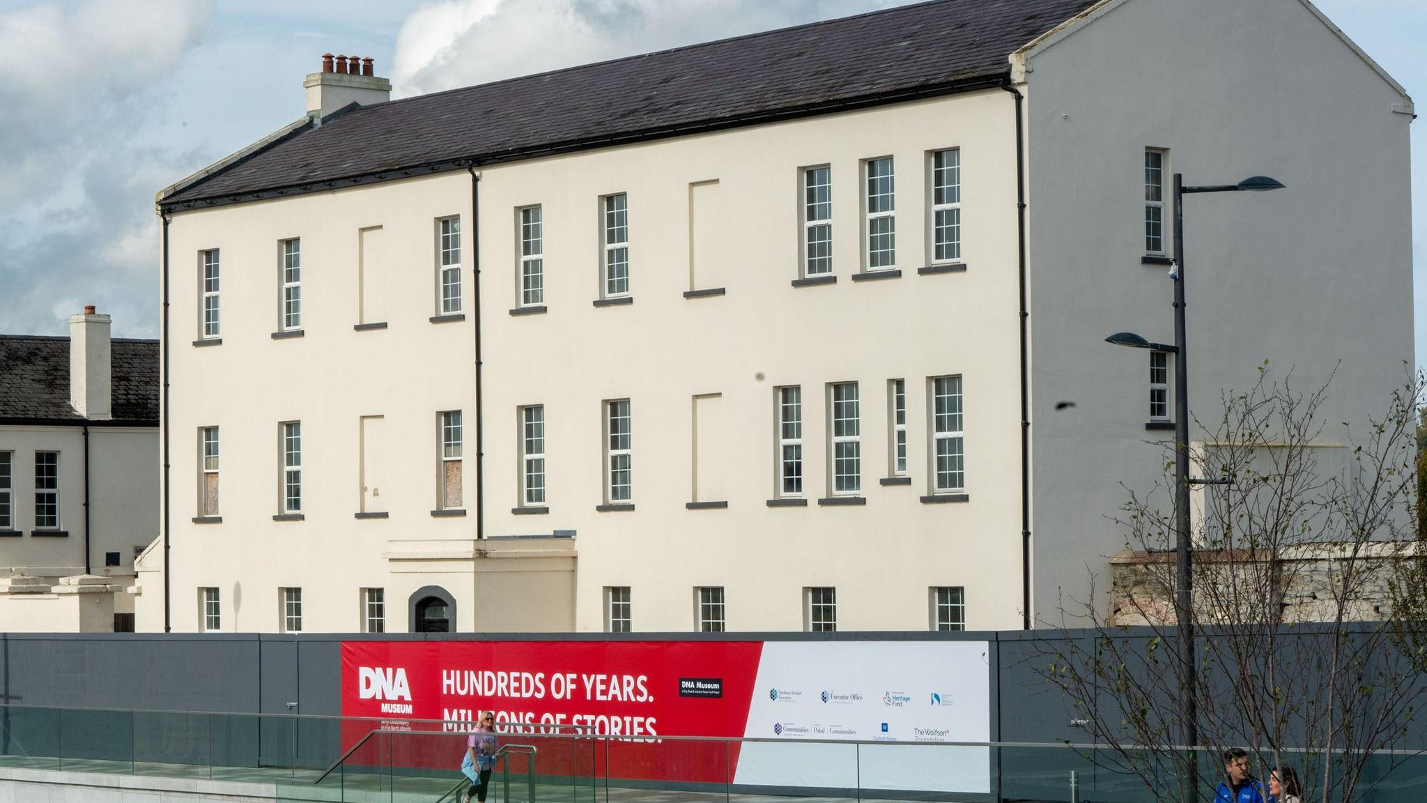 The image shows a large building, which is painted white, and set to house a new museum in Derry's Ebrington Square. It has three storeysand on its front is a series of windows. A porch can be seen at the front of the building, while a hoarding, with a sign for the DNA museum, runs along its outside. Three people are walking past the building.