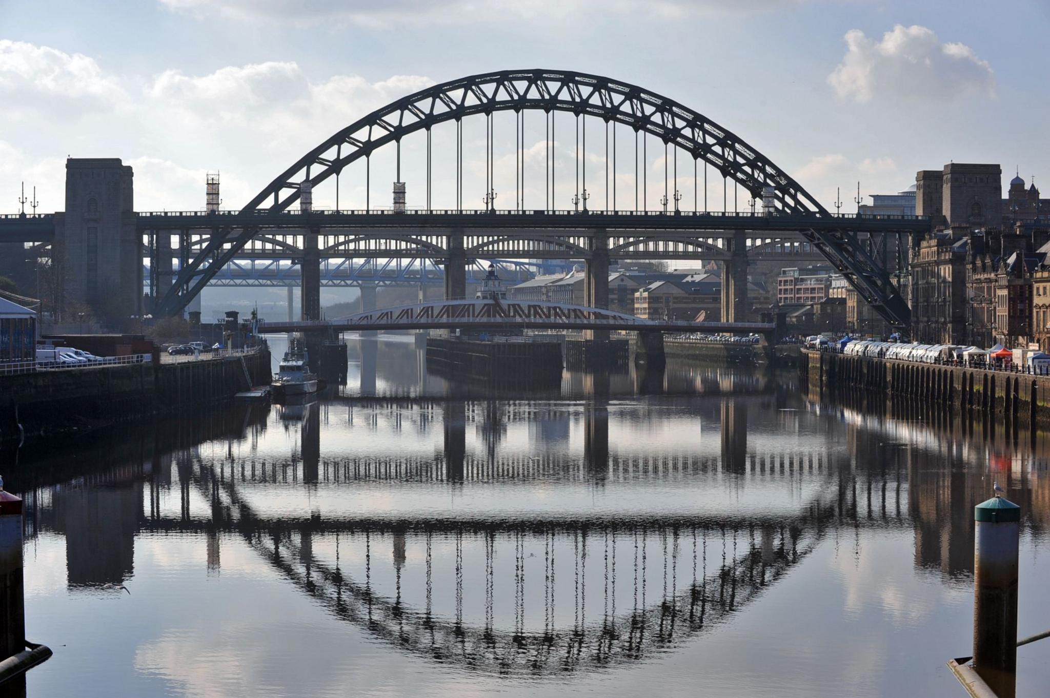 A wide view of the Tyne Bridge from the river Tyne. It is a metal bridge, painted green. Several other bridges can be seen behind it. The silhouettes of the bridges are reflected in the water below.