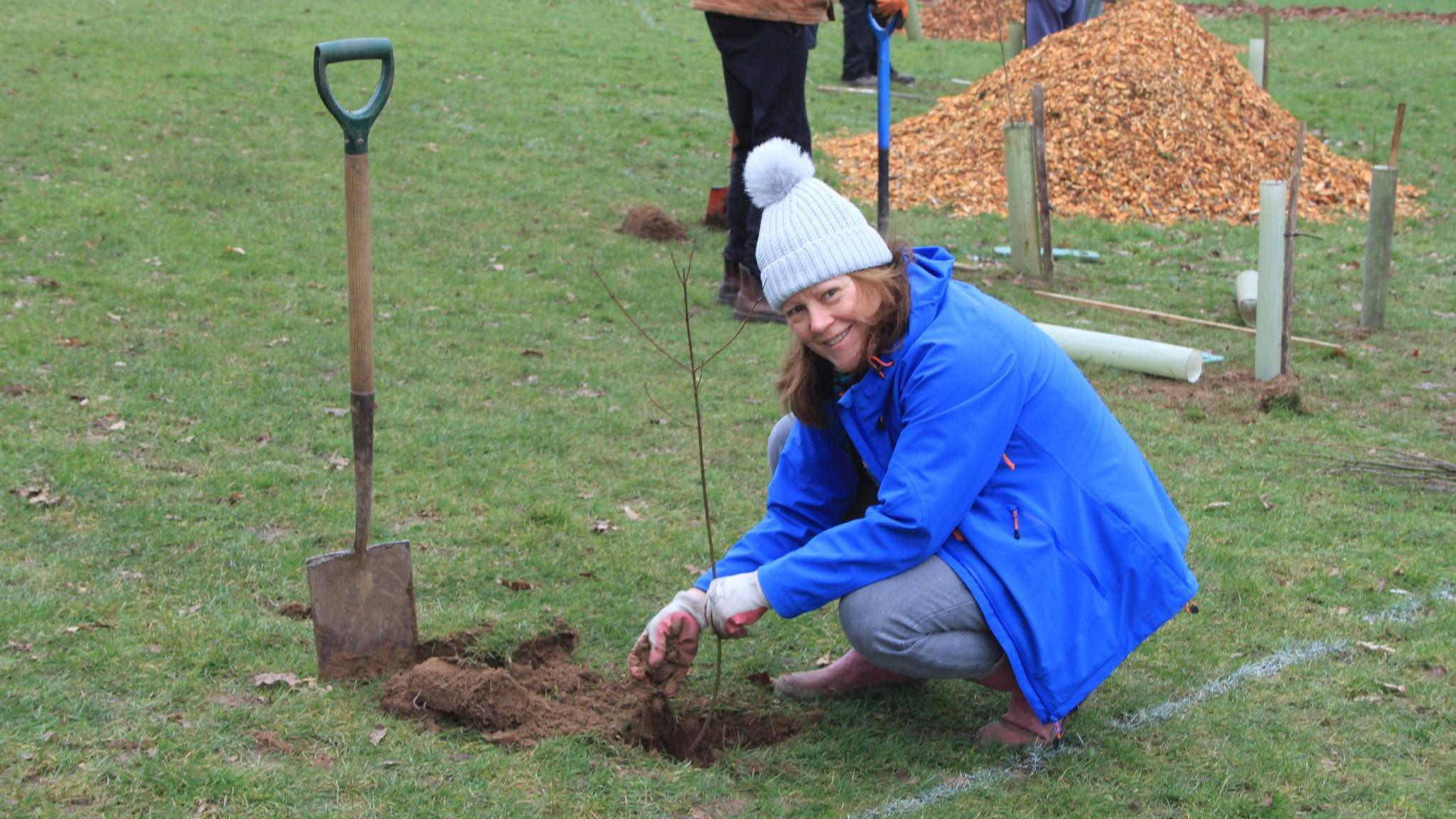 A woman wearing a blue jacket and a bobble hat planting a tree while looking into the camera