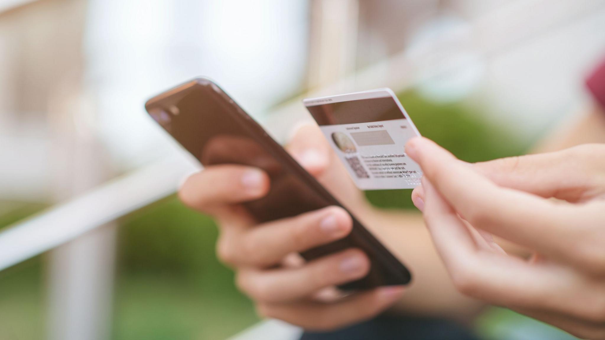 An anonymous teenager holding a bank card in their left hand and a mobile phone in their right