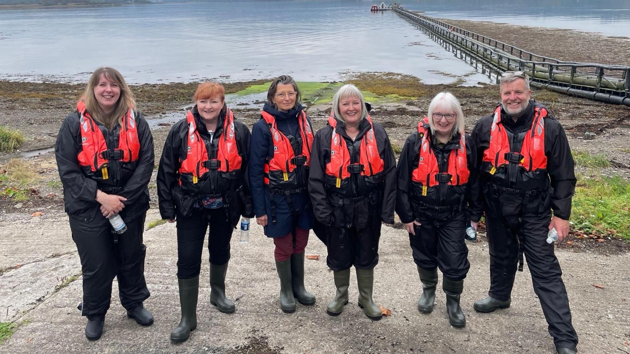 MSPs in life vests lined up waiting to take a boat trip to the Dunstaffnage fish farm on Monday. Left to right are Emma Roddick, Emma Harper, Ariane Burgess, Rhoda Grant, Beatrice Wishart and convener Finlay Carson. 