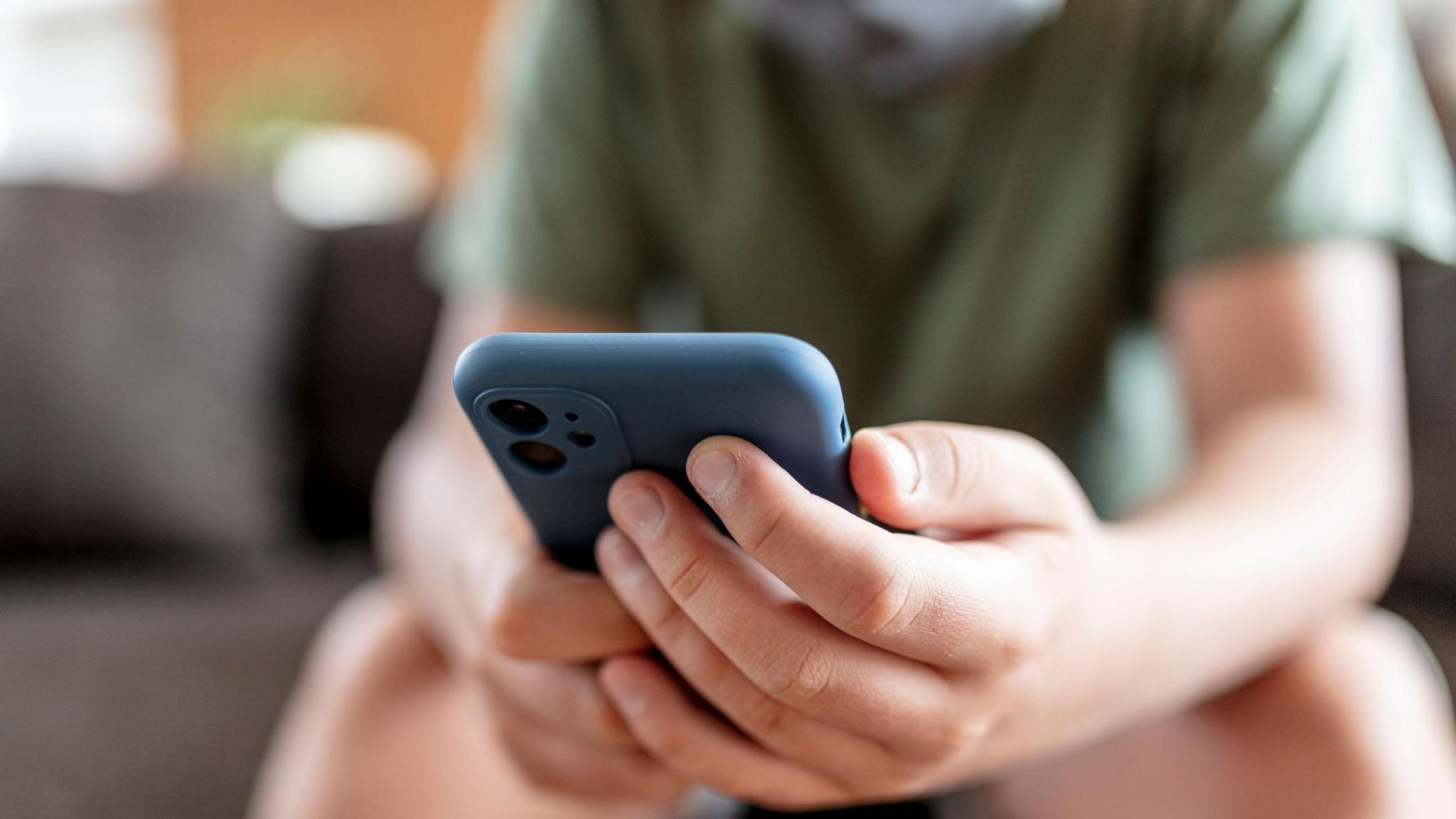 A teenage boy, wearing a dark green t-shirt, is shown blurred, sitting on a sofa. The image is focused on a blue smartphone he is holding in his hands.