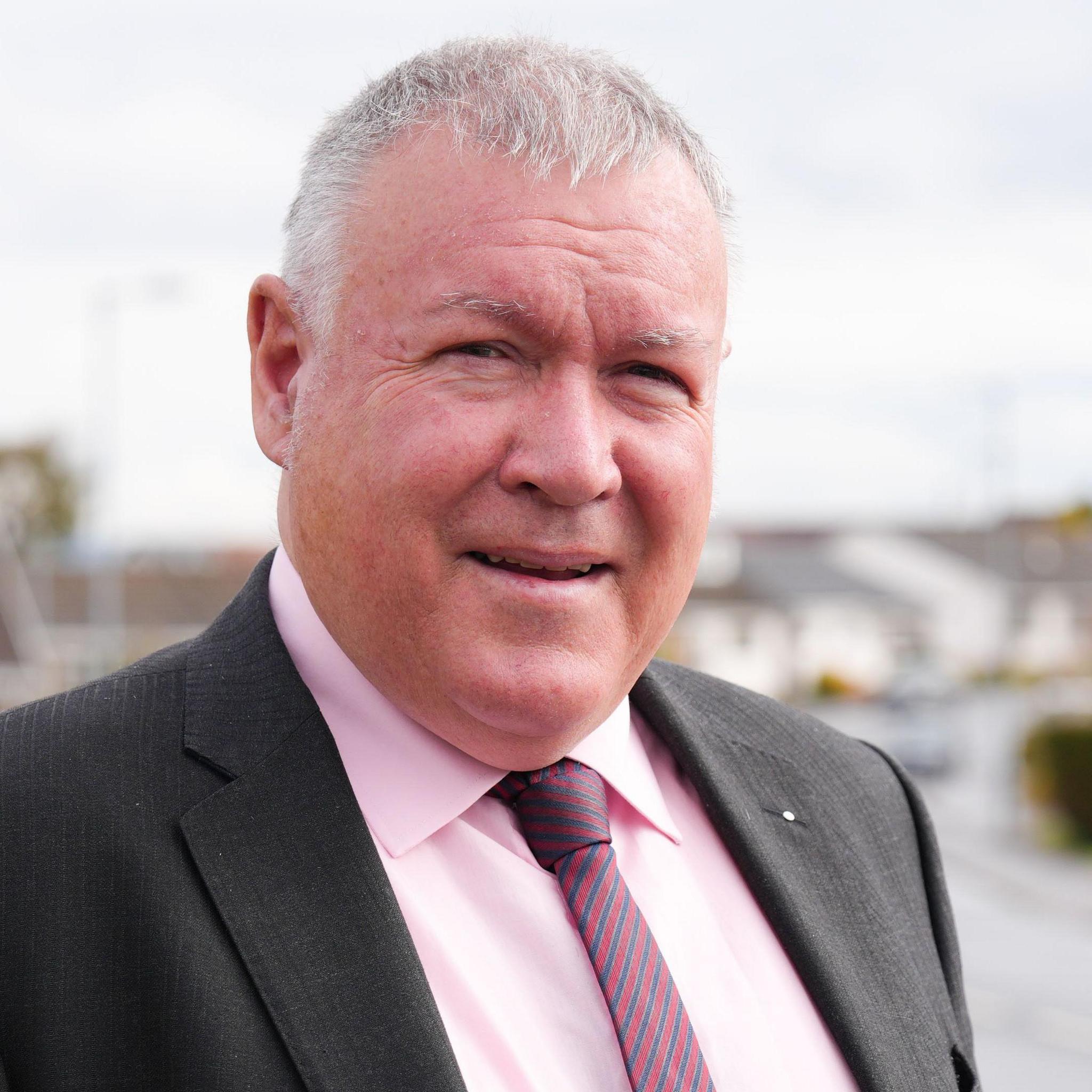 A man with short grey hair, wearing a dark coloured suit, a pink shirt and a striped tie. Houses can be seen in the background.