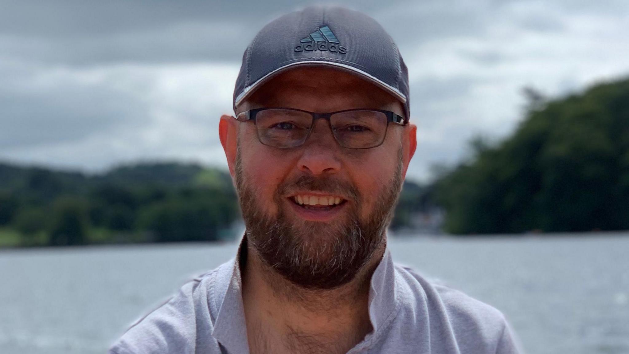 Andrew Slorance wearing a black adidas cap with a dark logo on the front, wearing an open grey polo shirt with the collar pulled up in front of a body of water and two areas of greenery under a grey sky.