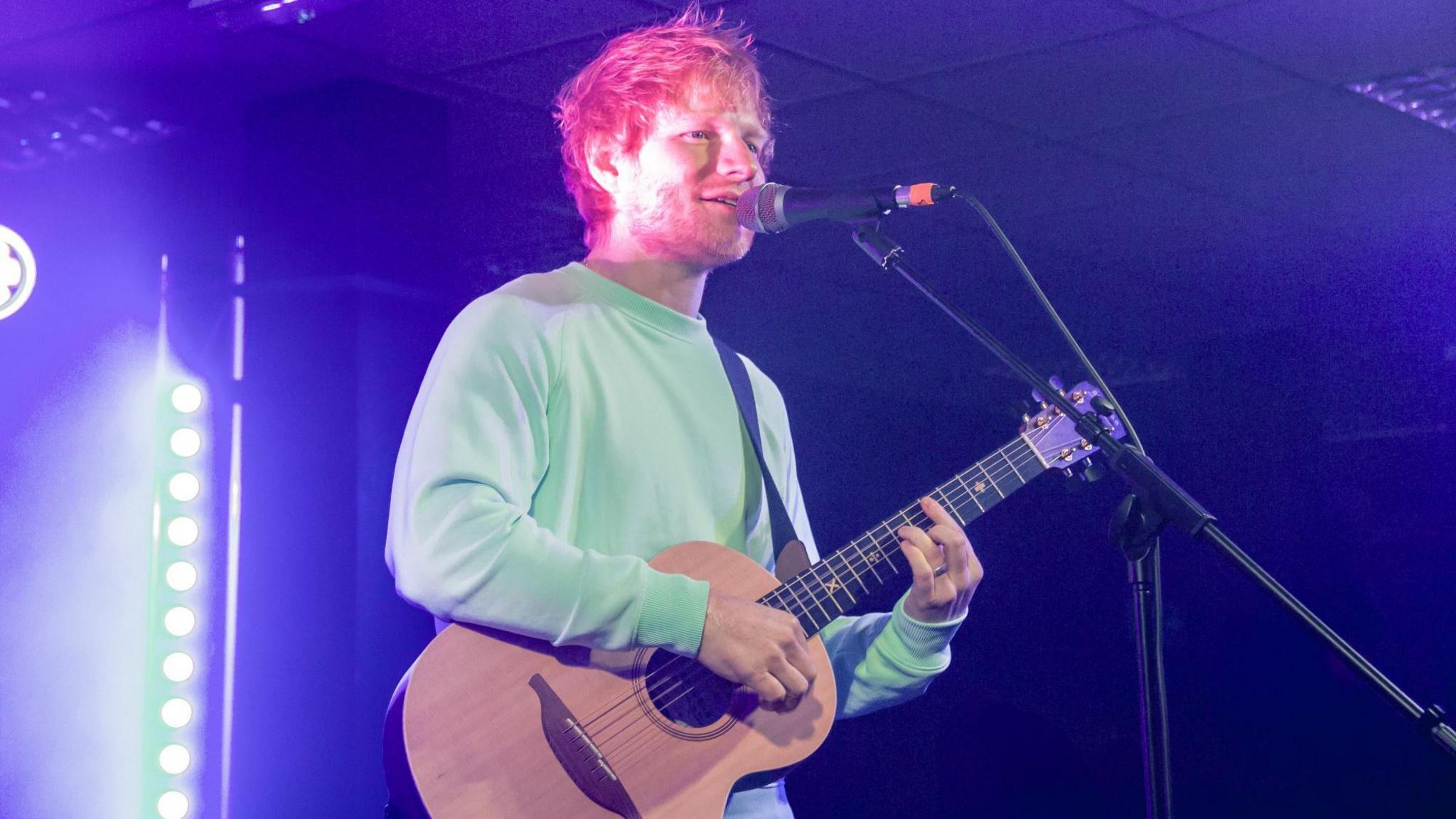 Man with a ginger hair and beard holds a guitar and stands before a microphone. He is strumming the guitar and is wearing a pale green sweatshirt. He is on a stage and is lit from behind in a darkened room.