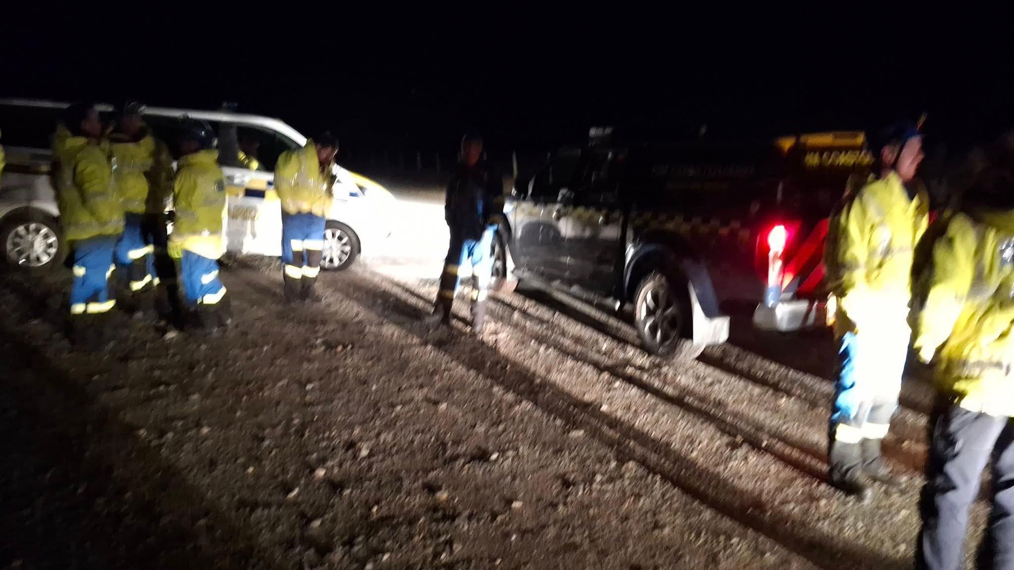 A slightly blurry picture of the search and rescue team which is made up of around seven people and they are all dressed in yellow jackets and blue trousers. There are two vehicles parked on some gravel behind them and it is dark.
