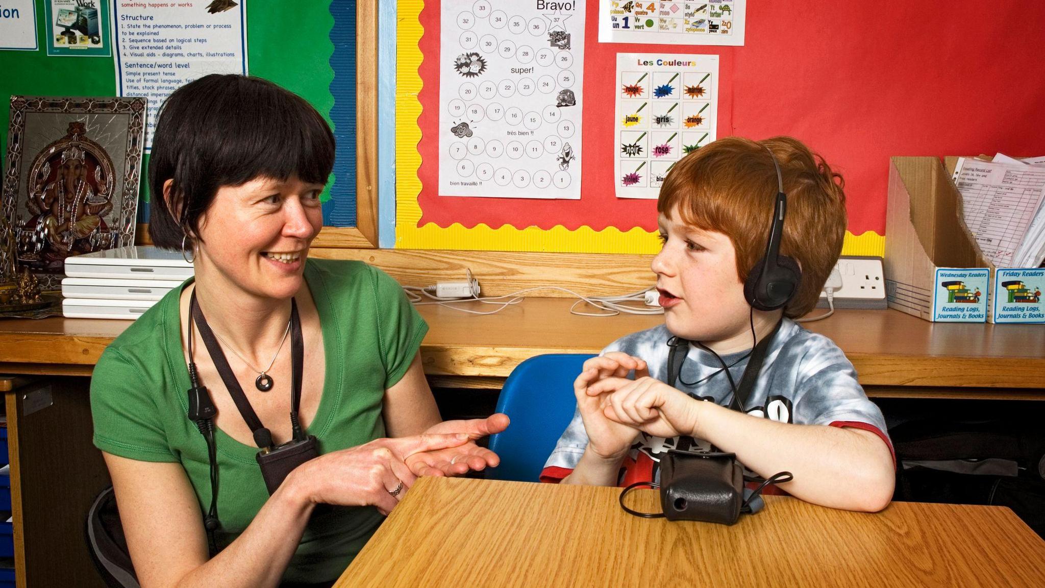 A deaf school boy in a school class room using sign language. An seven year old red head boy fitted with a voice amplifier communicates with his teacher using sign language.