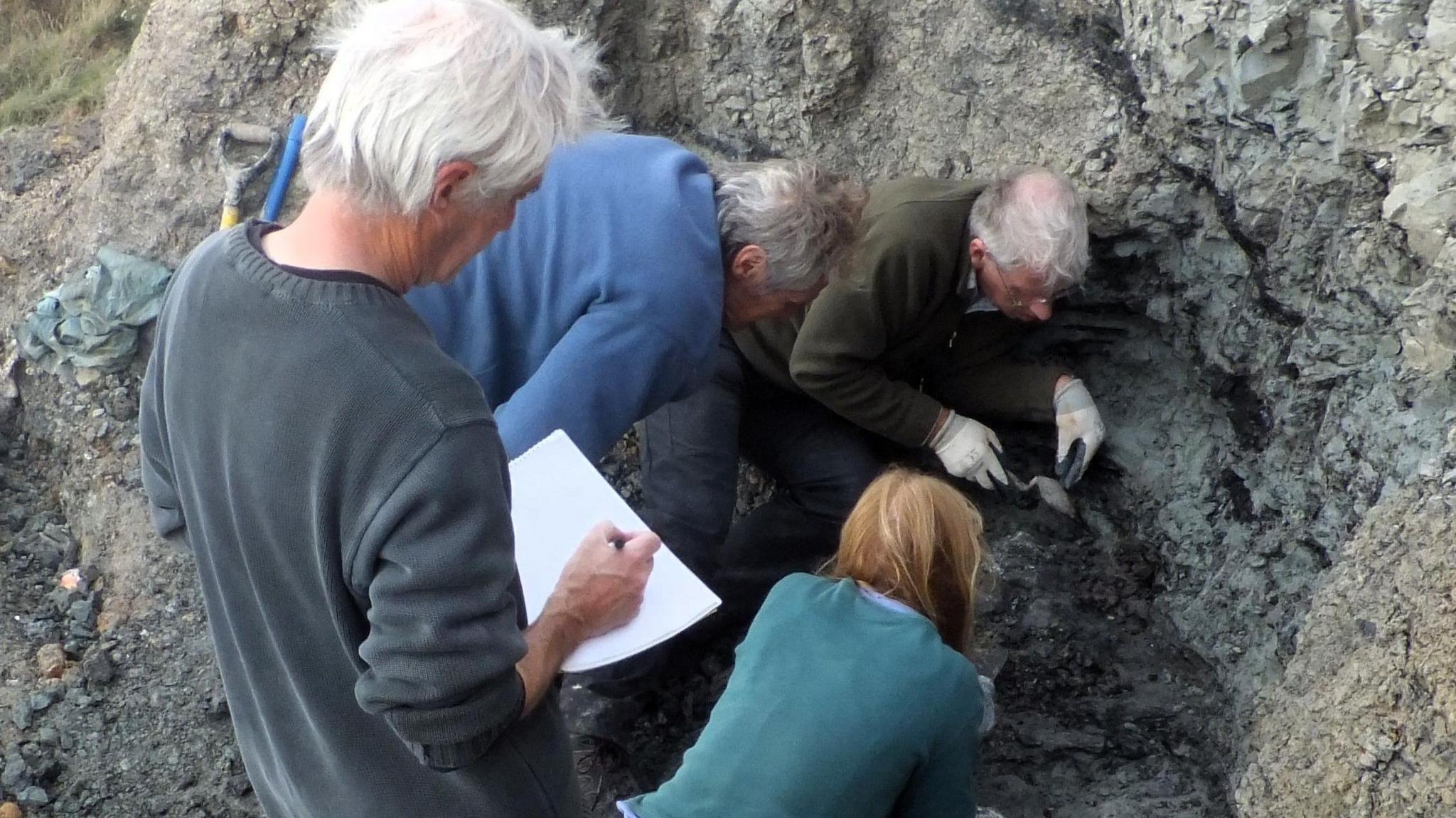 Rear view of Nick Chase sketching in an A4 book. He stands above three people who are peering into a hole dug into the grey cliff.