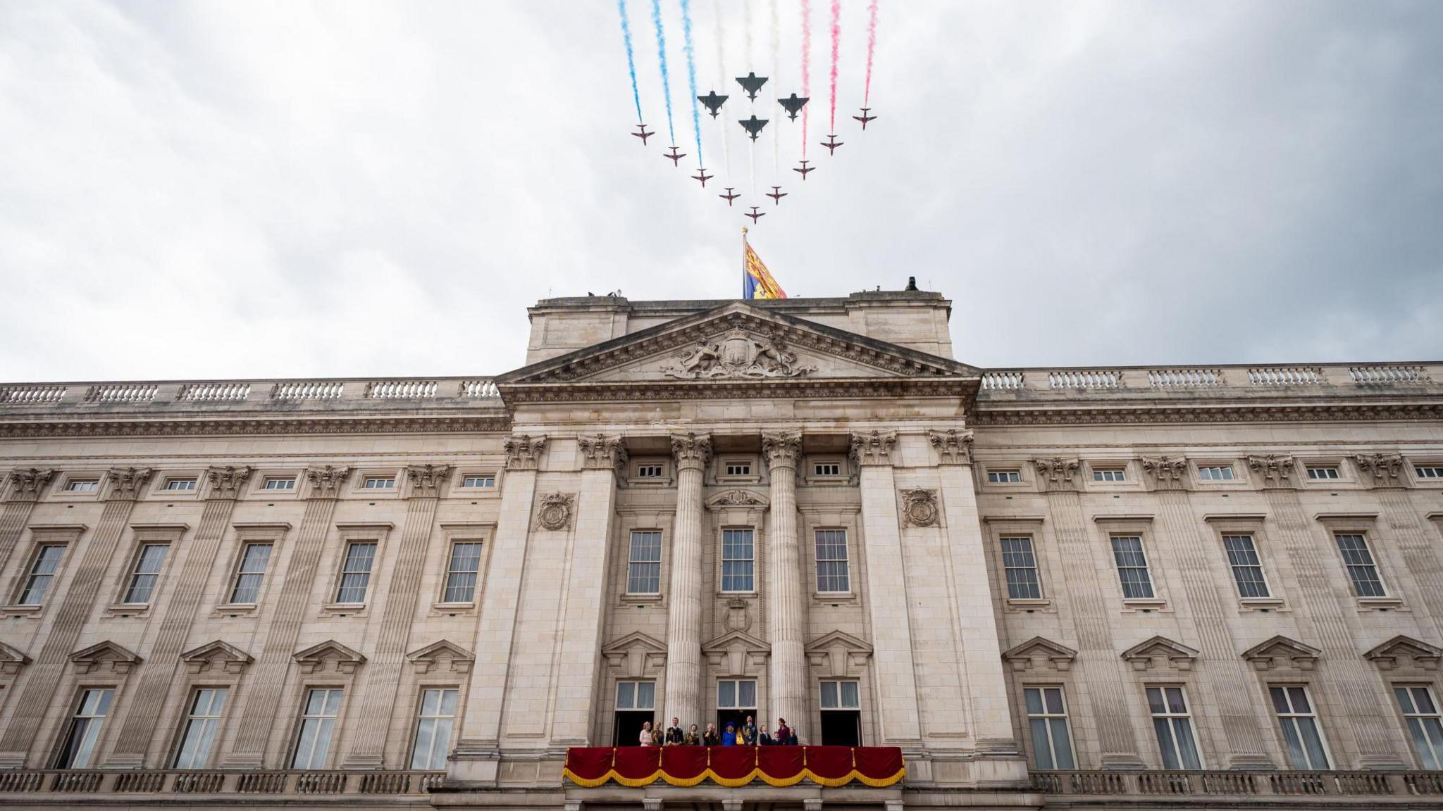 Red Arrow planes and fighter jets fly over Buckingham Palace. The Royal Family are on the balcony, which is draped with red cloth.