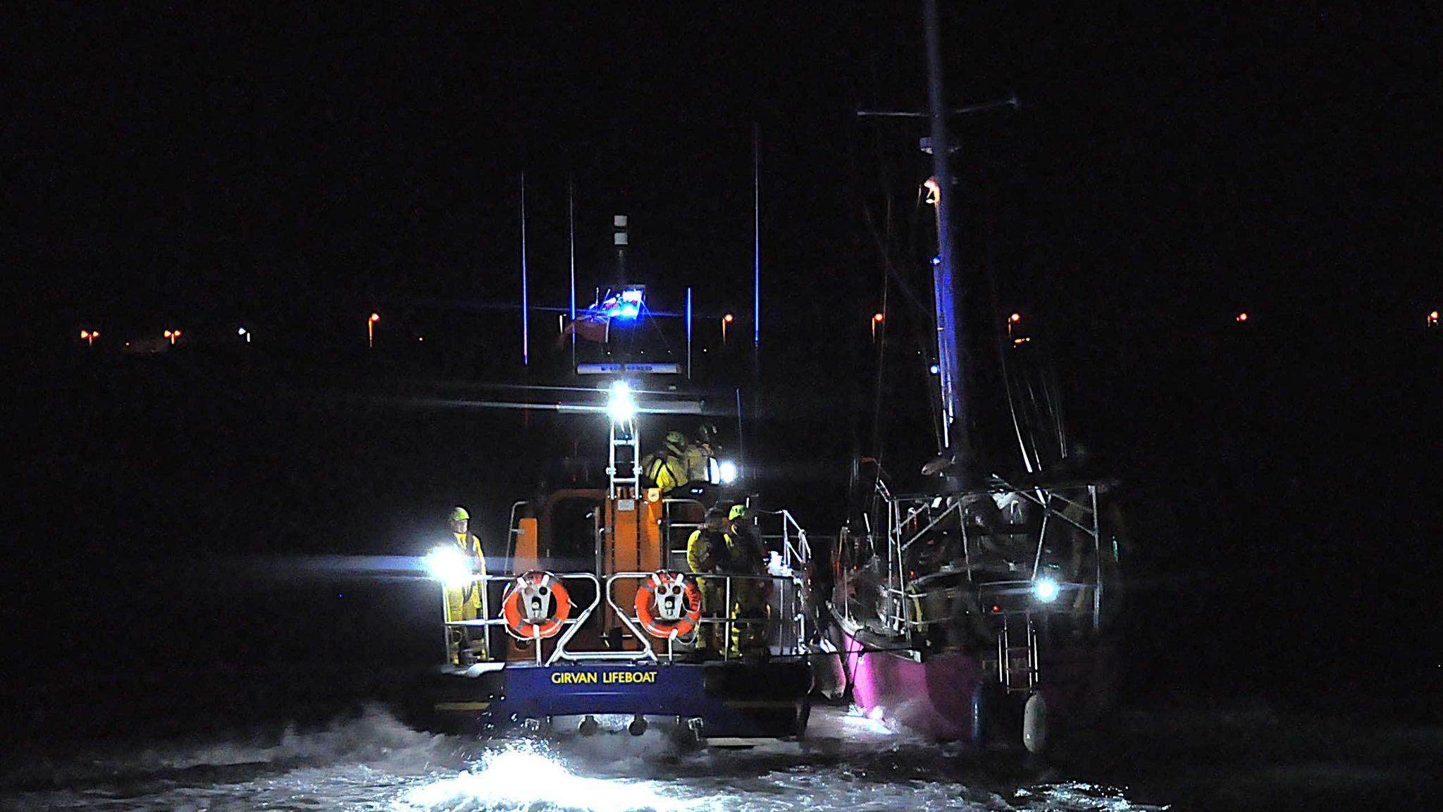 A dark image of a rescue operation with the Girvan lifeboat and three or four crew in yellow gear aboard towing a pink yacht to safety.