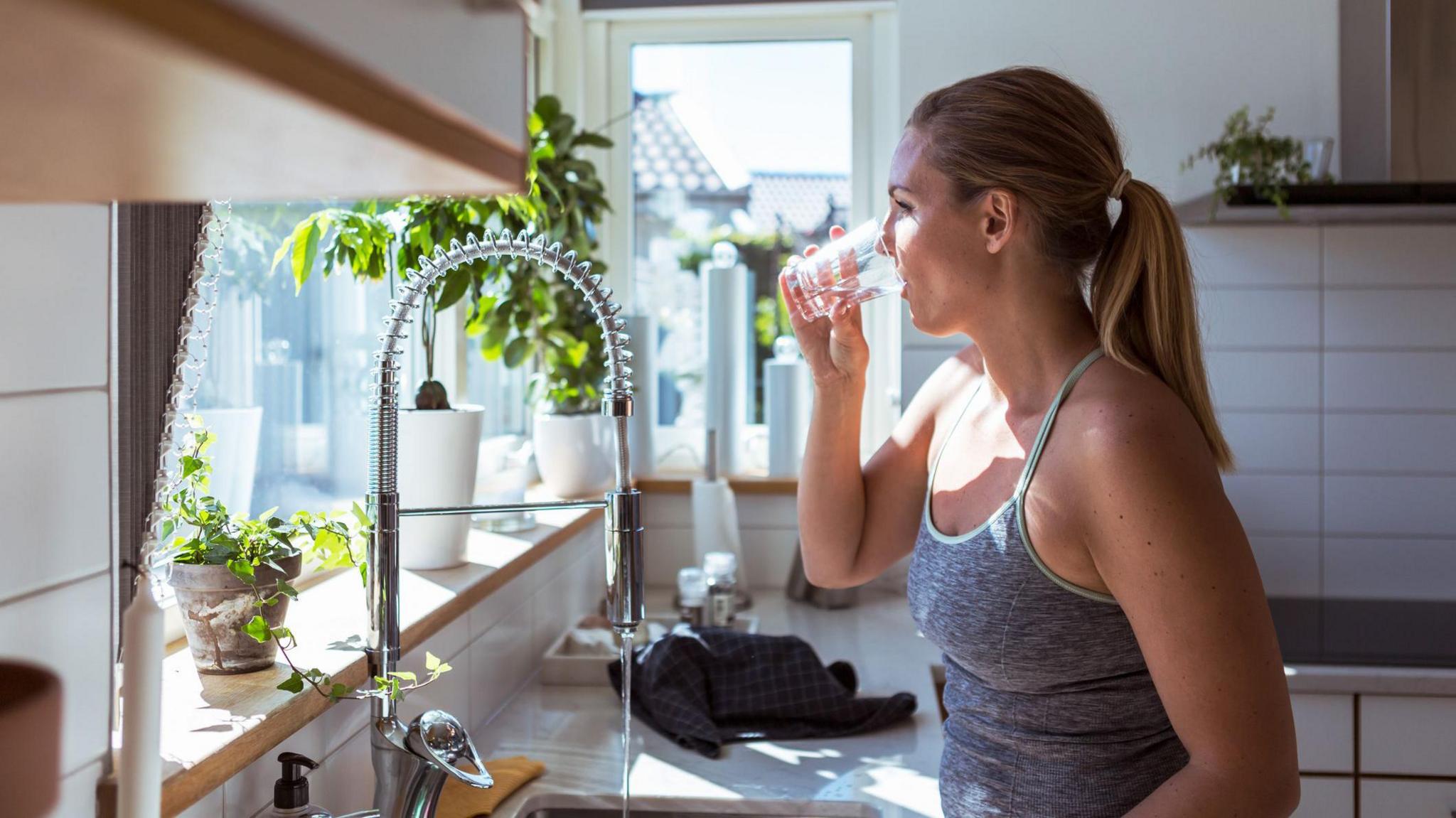 A woman in a vest top with blonde hair tied at the back drinks a glass of water standing by a kitchen sink. The white-tiled kitchen has pots of green plants on the window sill and has a big glass window allowing sunlight into the kitchen. 