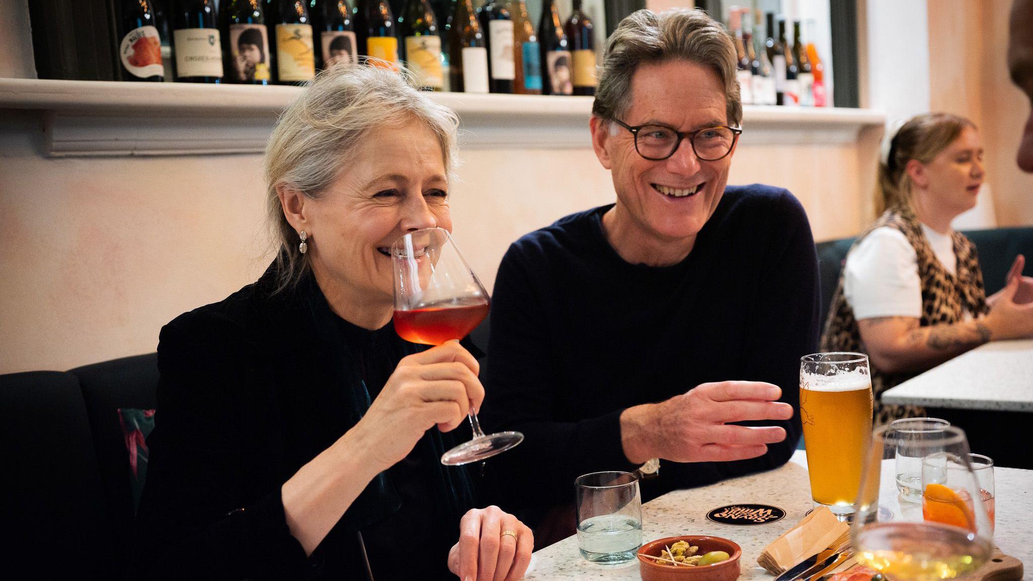 A man and a woman smile as they sit at a table inside the KASK wine bar on Cheltenham Road in Bristol. They are both wearing dark tops and she has a wine glass to her lips. He has a pint of beer in front of him. The wall behind them is a neutral colour with one shelf on it which has a row of wine bottles on it.