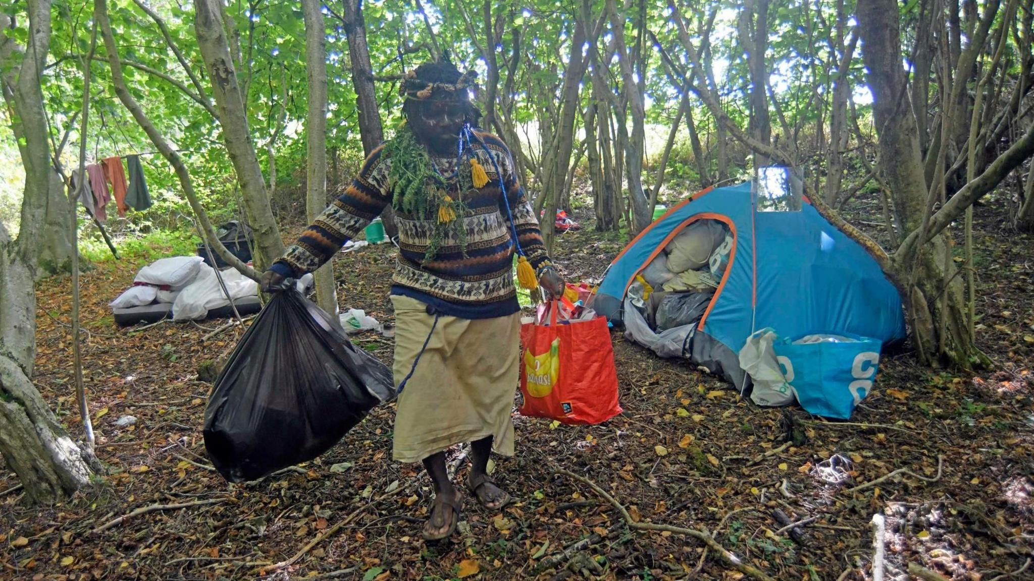 Kofi Offeh walking beside a makeshift camp in the woods. He is carrying a black bin bag and a bag for life with items inside. There is a blue tent crammed with items such as bedding and toilet roll and there are a variety of bags and other items scattered abot. There is a washing line with clothes hanging on it between some trees.