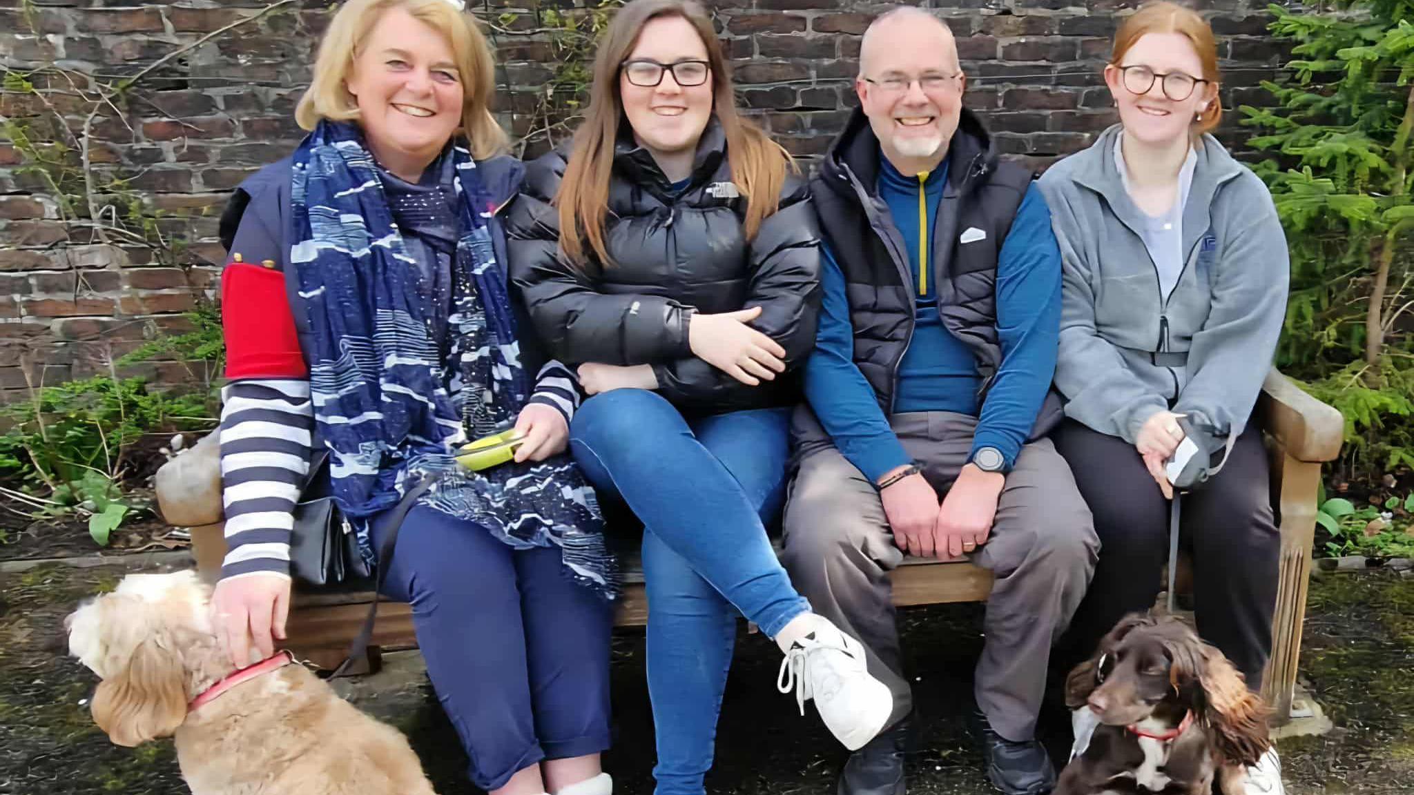 A family of four sat on a bench with their two dogs at either side. There is a woman, a teenage girl, a man and another teenage girl.