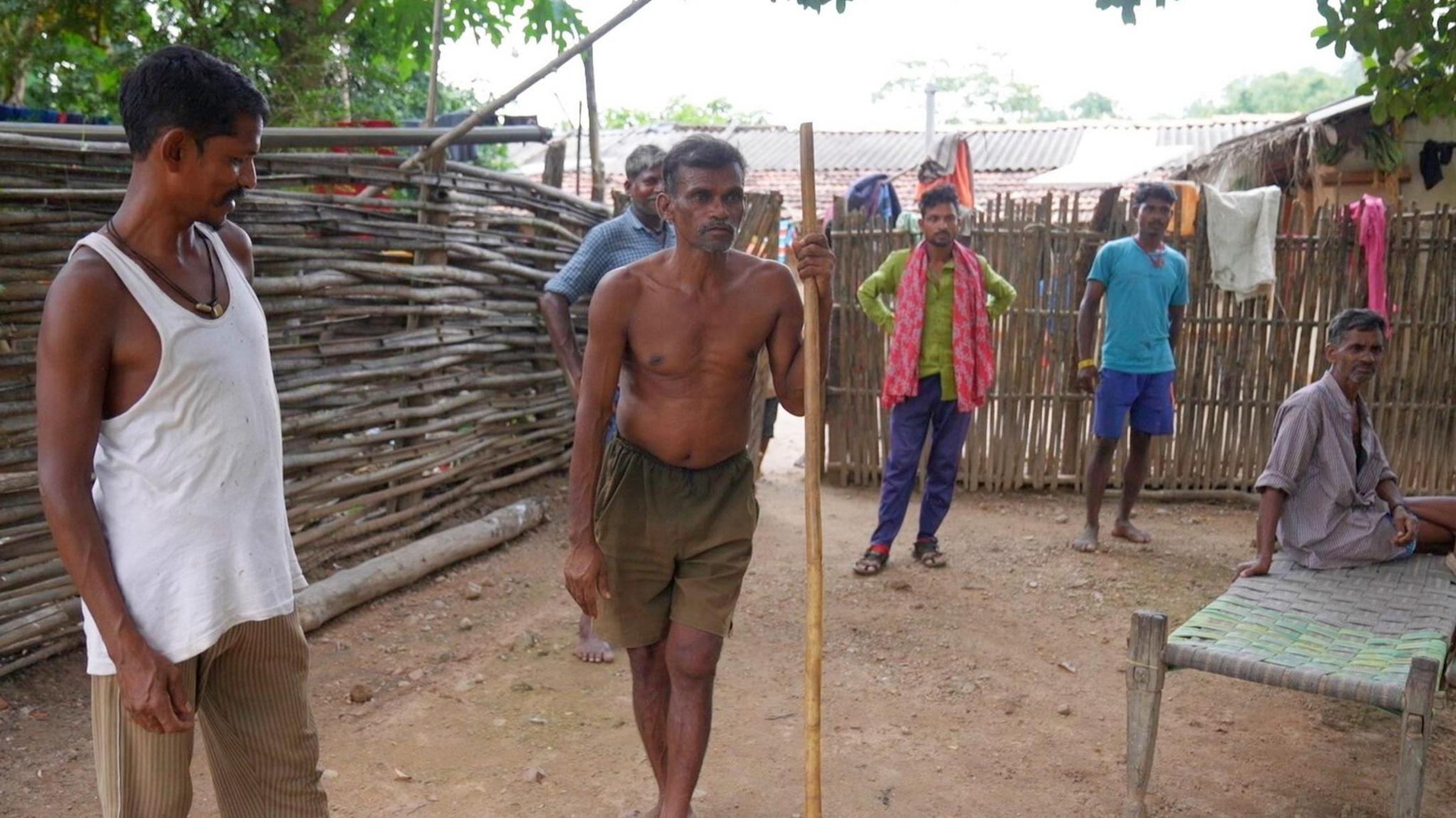 Men standing in a compound in Bastar, Chhattisgarh. The man in the centre is wearing only shorts and is holding a stick while others stand on the sidelines. 