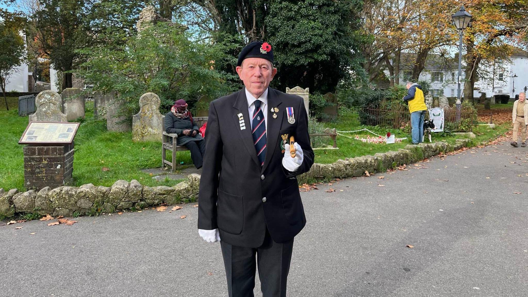 Tony Avery wearing a dark blazer, service medals and beret, standing to attention in front of Shoreham's war memorial.