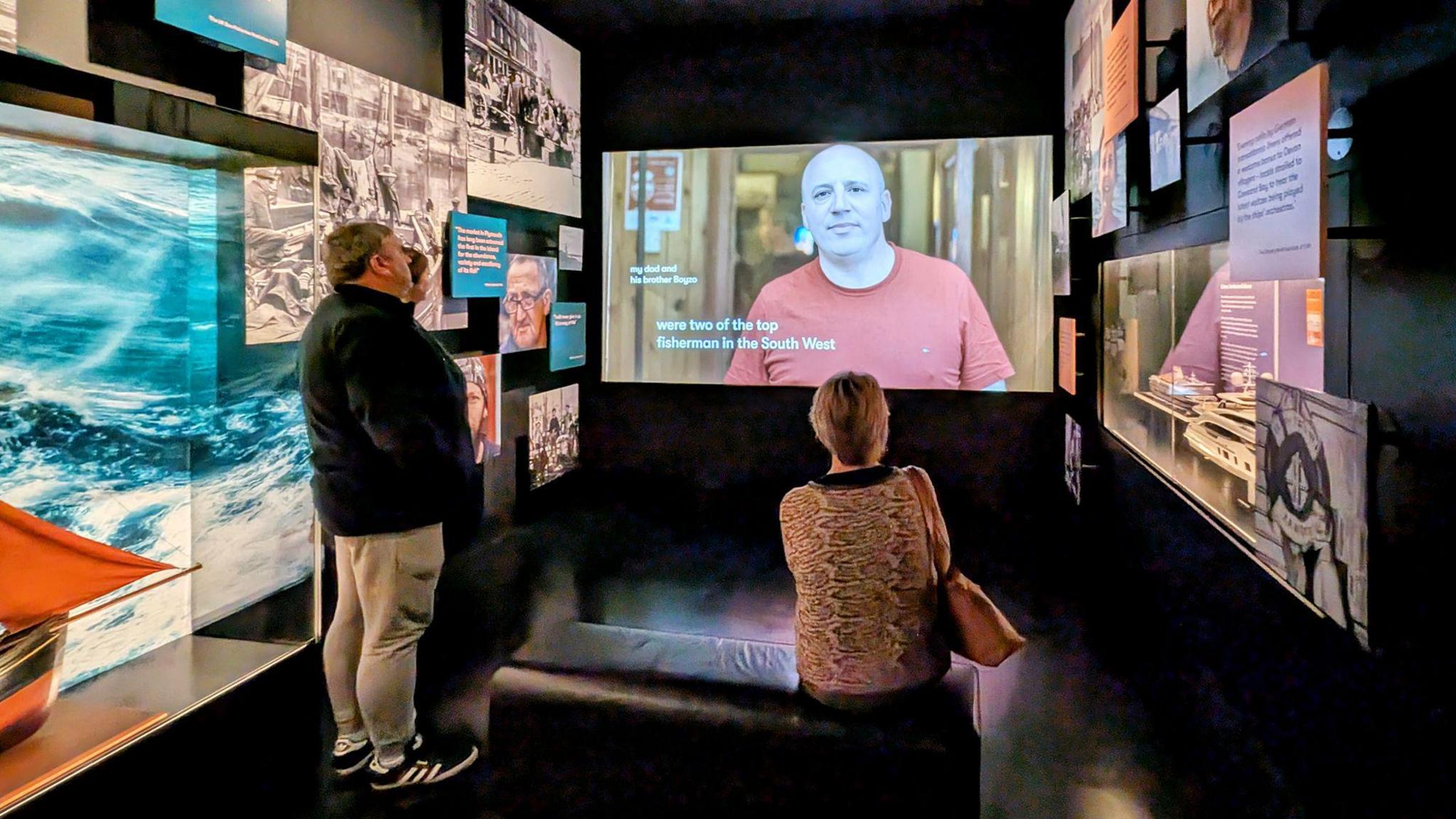 A man is standing and a woman is sitting on a black leather bench and they are watching a video screen which shows a man and the text 'We're two of the top fishermen in the South West'. It is a small square room and the walls are covered with photographs of the sea and boats and there are panels covered in text.