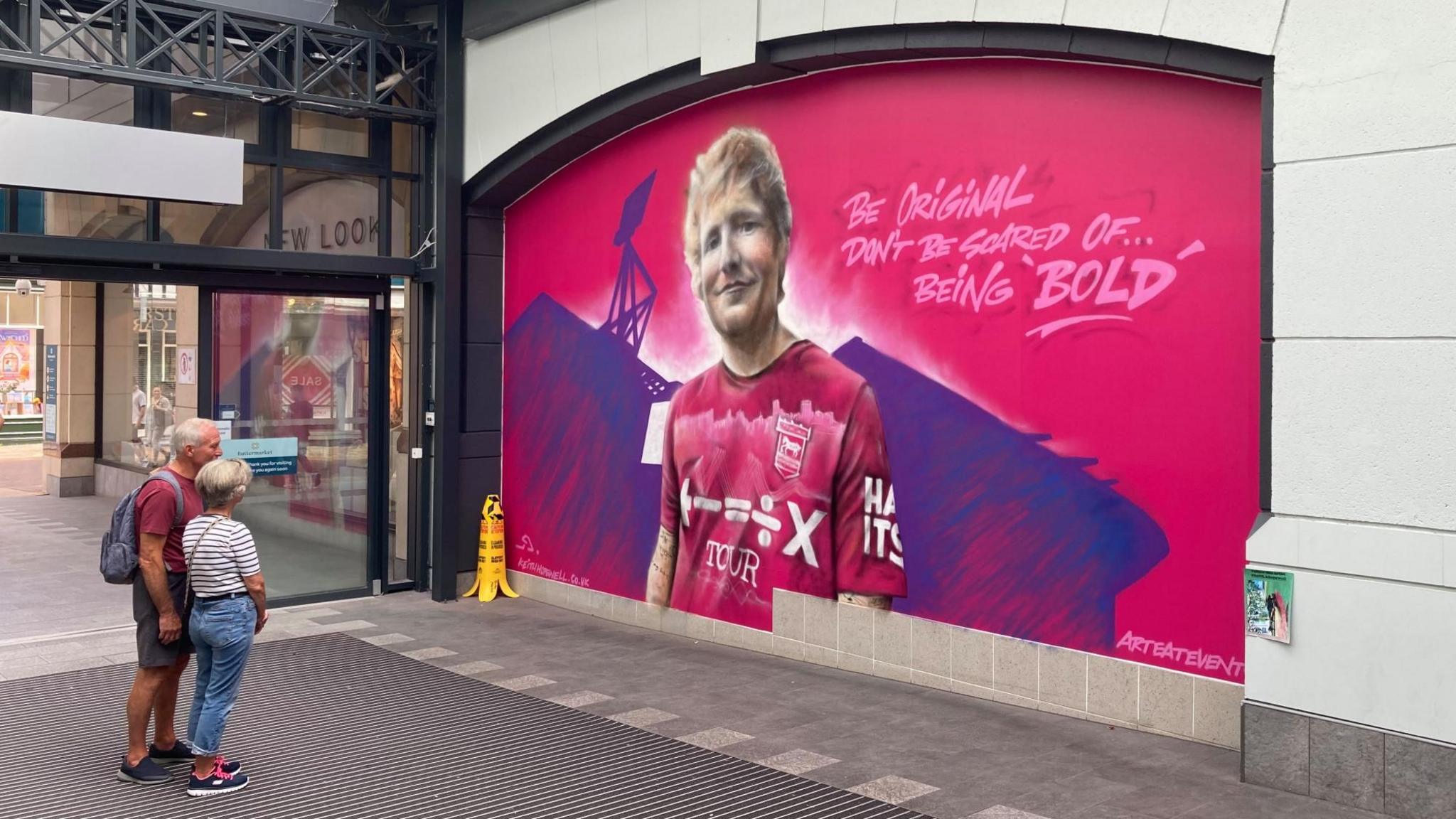 A mural of Ed Sheeran wearing Ipswich Town's pink alternative shirt with the words "BE ORIGINAL DON'T BE SCARED OF... BEING 'BOLD'" written to the left of his head. A silhouette of Portman Road football ground is behind him. A couple look at the picture which is just inside the entrance doors to a shopping centre.