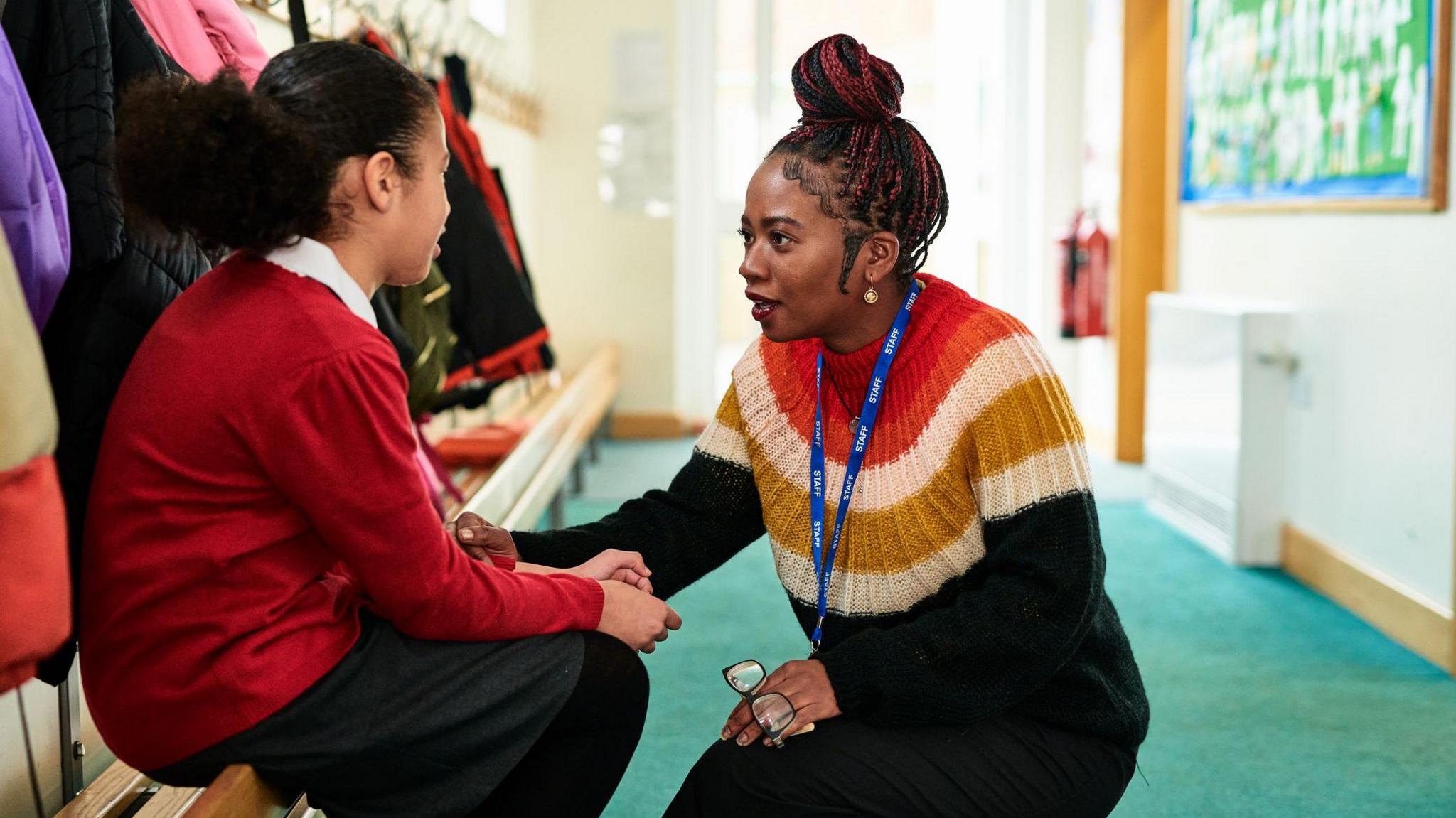 A primary school student in a red jumper sits on a bench being comforted by a school teacher.