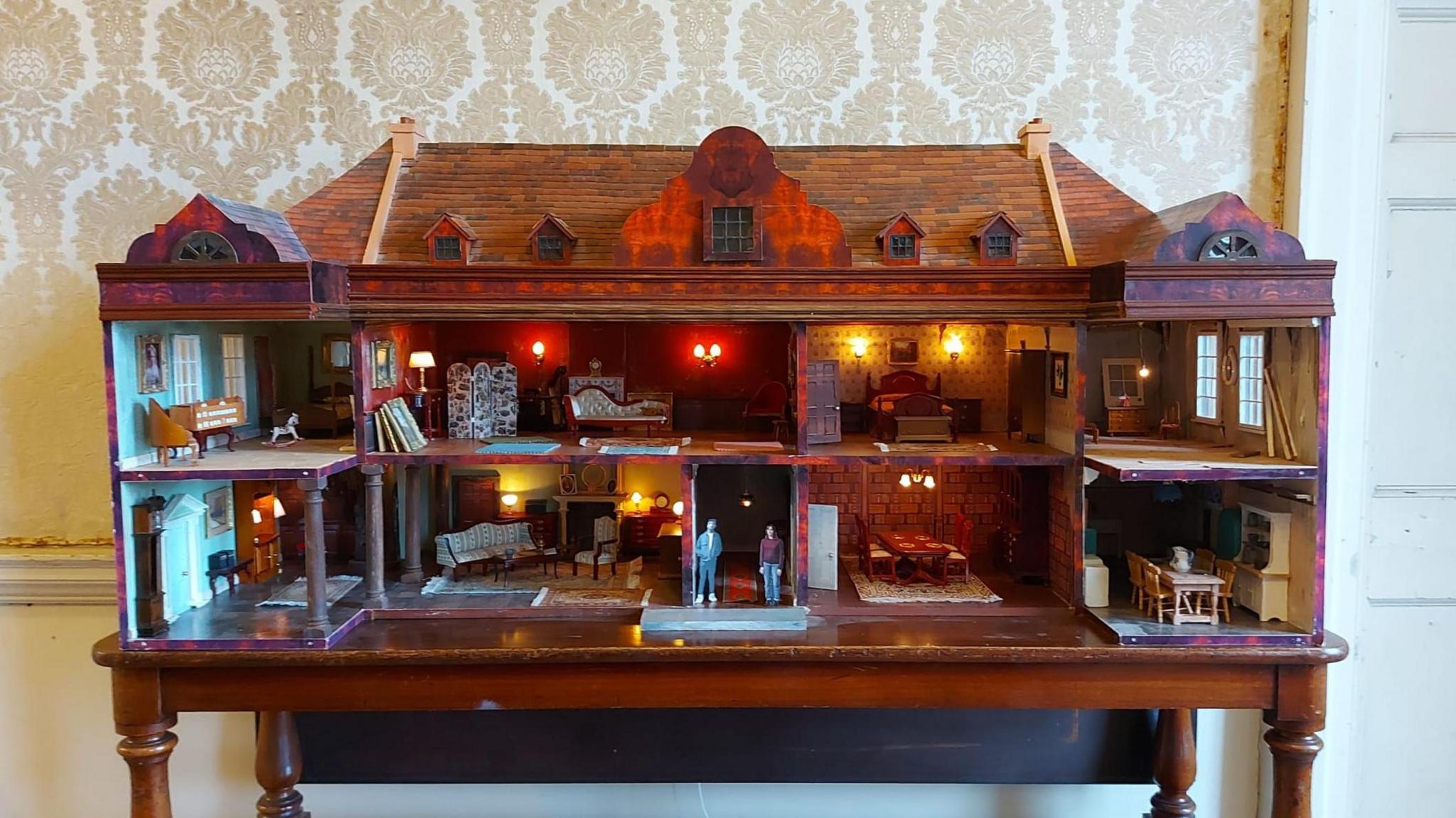 A dolls house version of West Horsley Place, opened to show the interiors details of the house, with models of the two living characters from the show placed in the entrance.