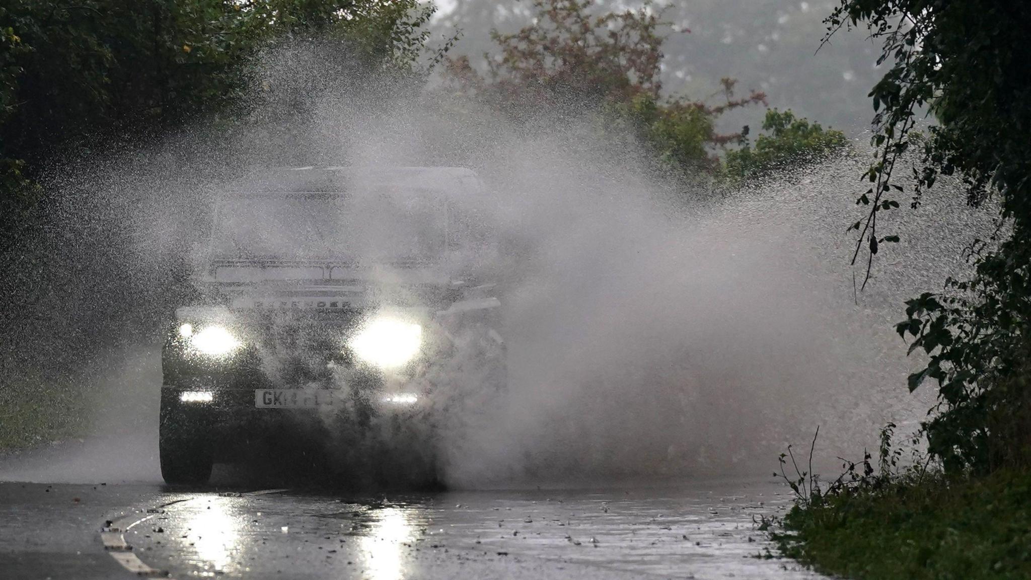 A jeep-style vehicle causes a huge wall of spray to fly up as it drives on a flooded road near Gloucester.