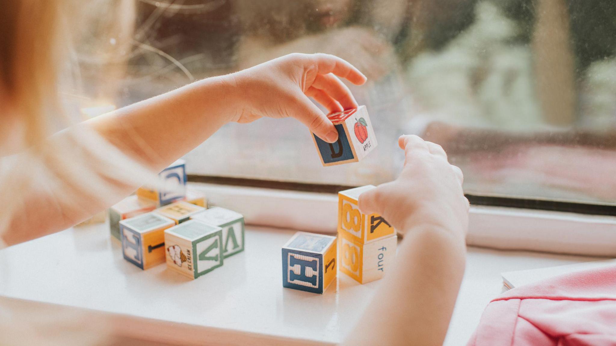 A close-up of a child's hands as they play with building blocks on a window sill.